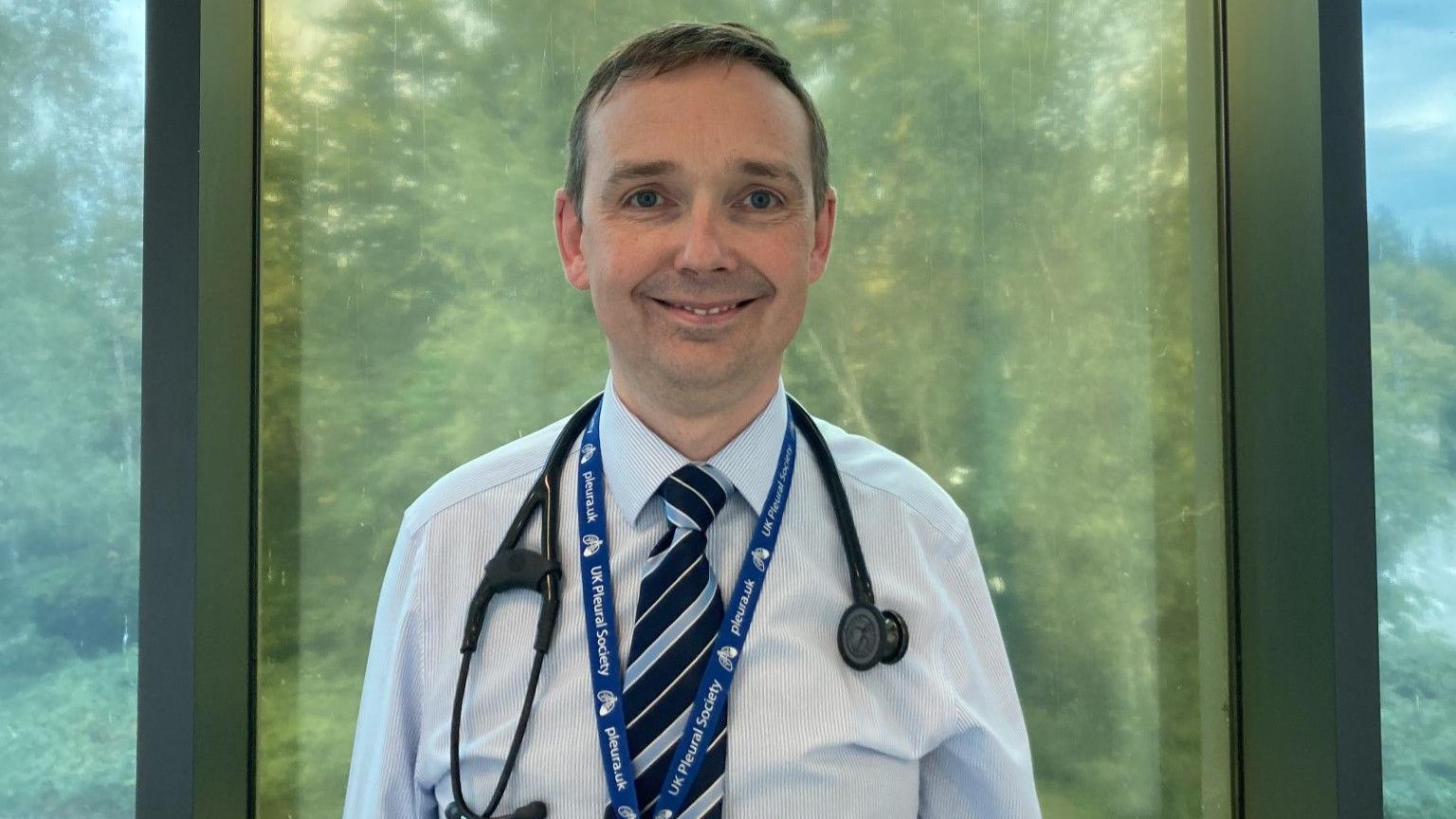Dr Mark Roberts is a respiratory consultant. He is wearing a stethoscope around his neck and a pale blue shirt and tie