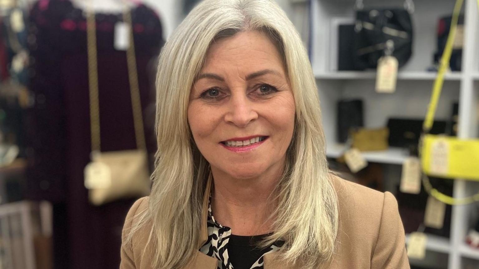 Helen Rutter with long blonde hair is wearing a yellow jacket. There are items on shelves in the background.