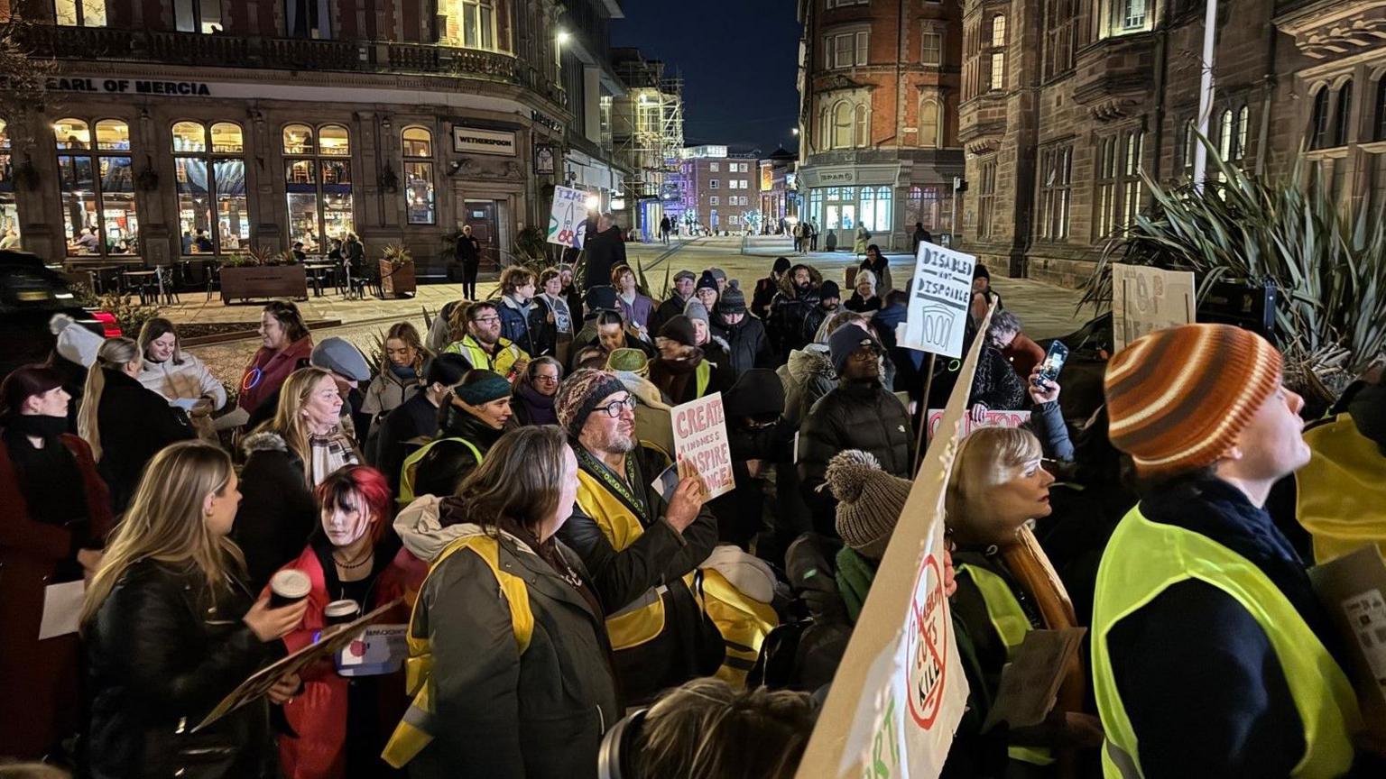 People holding banners protesting in the evening. Buildings are in the background.