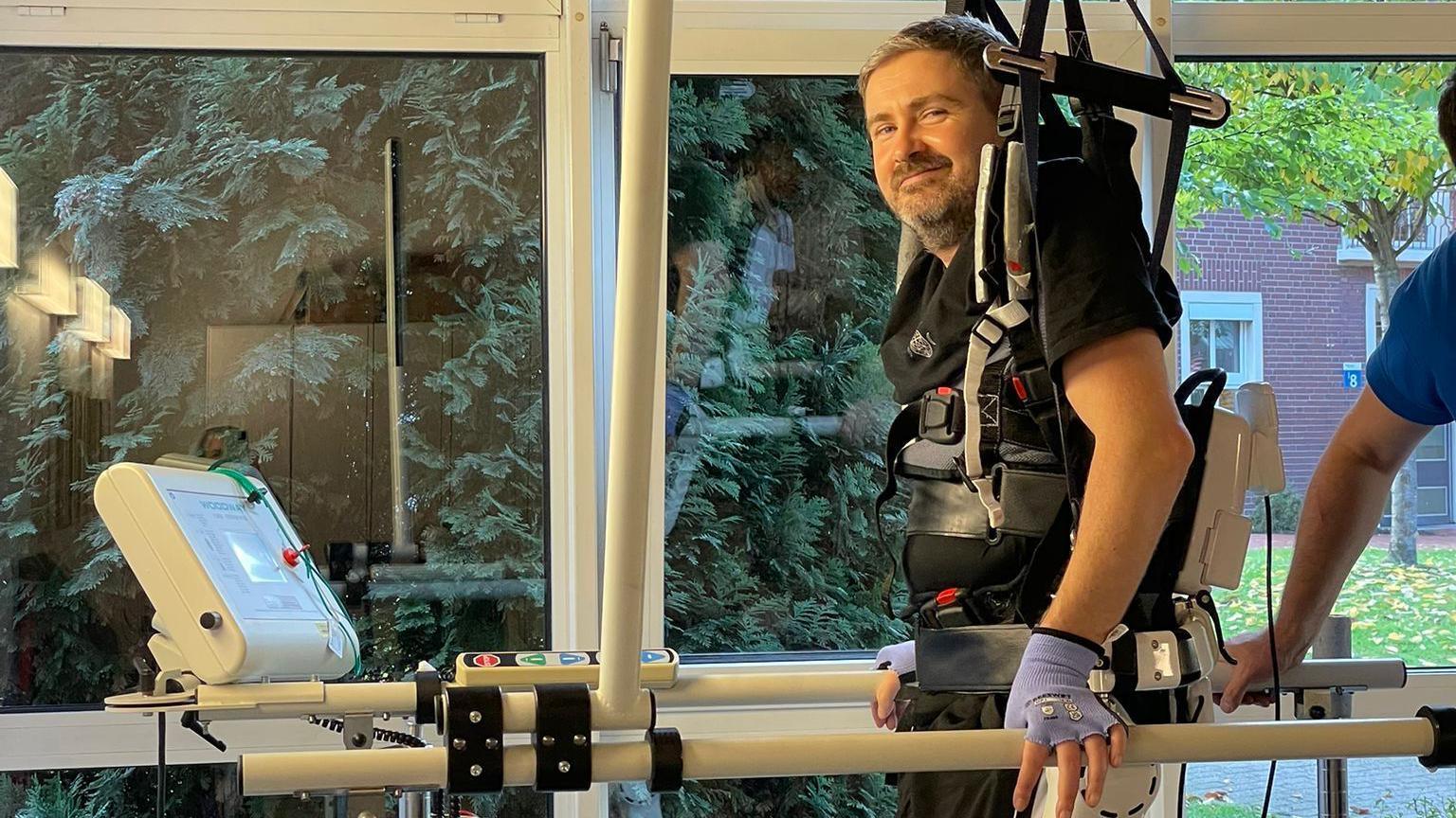 Dan Richards yn defnyddio peiriant i gerdded. Mae'n sefyll ac yn gwenu.