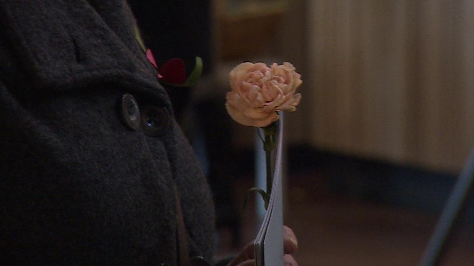 A pale pink flower is held by a woman, wearing a dark grey coat ready to lay at the church ceremony