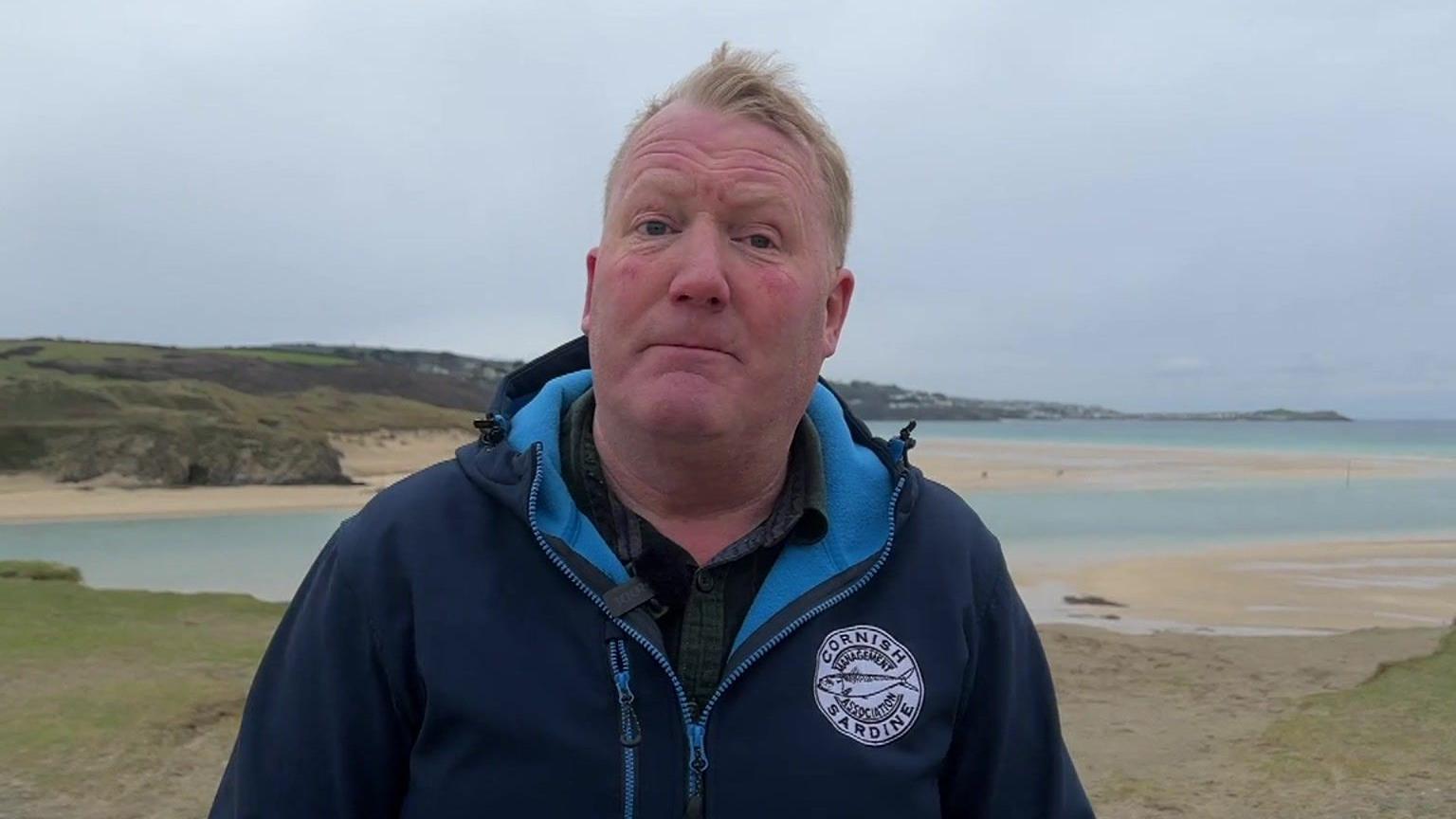 Gus Caslake stands in front of a bay along the Cornish coast while wearing a navy blue coat with a Cornish Sardine Management Association badge on it. Strips of sand are in the distance along with cliff edges and patches of grass. It is an overcast day with thick cloud cover.