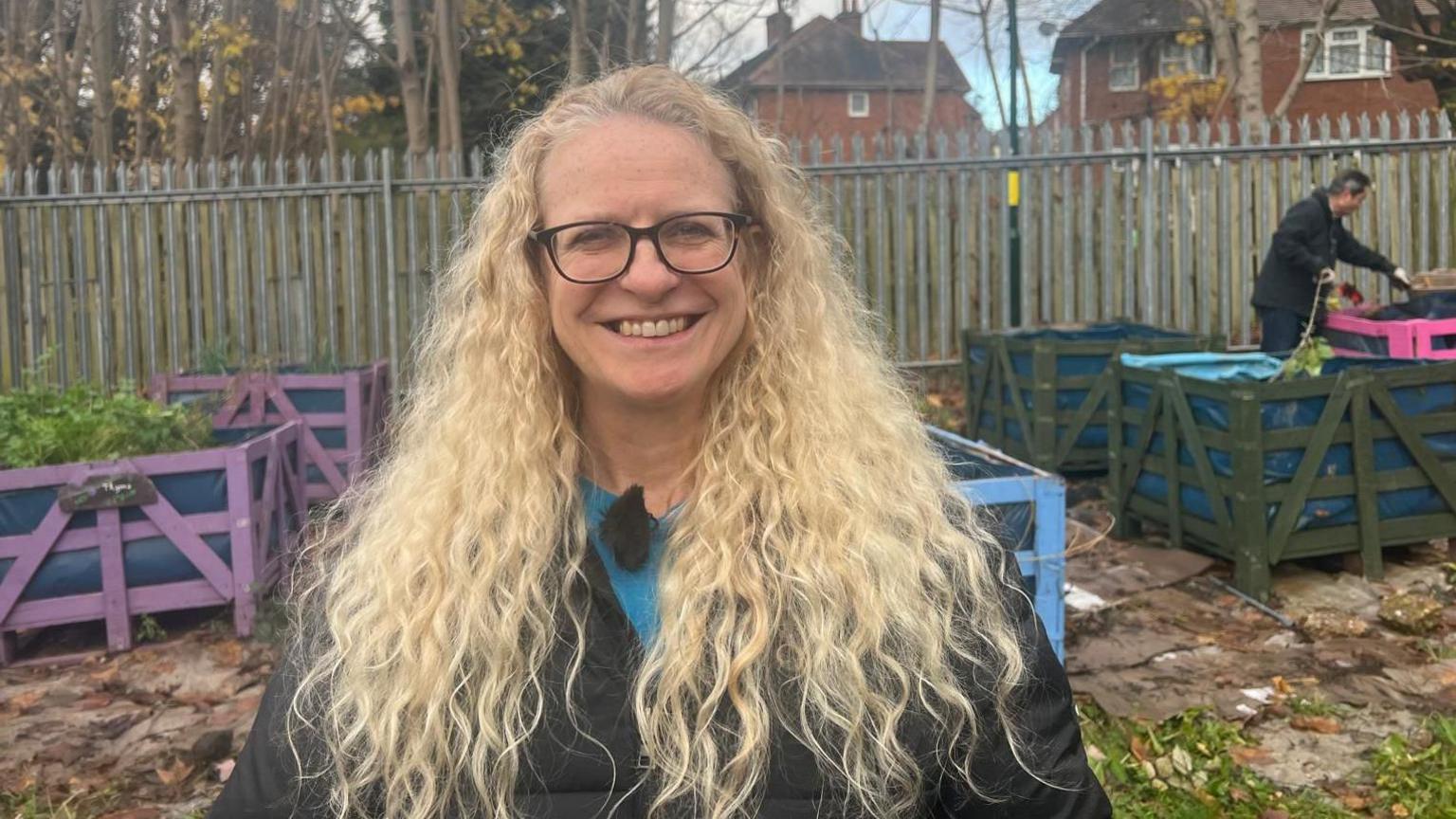 A woman with long blonde wavy hair and glasses is smiling and looking directly at the camera. She is wearing a black coat with a blue tshirt that is just visible below her neck