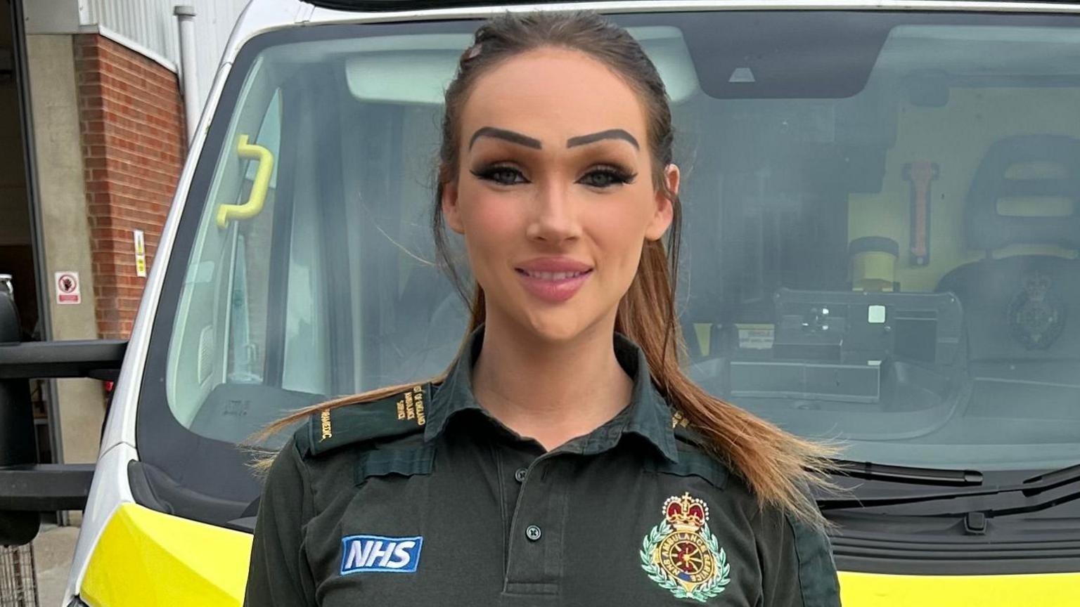 Phoebe Grace is wearing a dark green polo shirt, branded with the East of England Ambulance Service emblem. She has long brown hair in a ponytail and is standing in front of an ambulance.