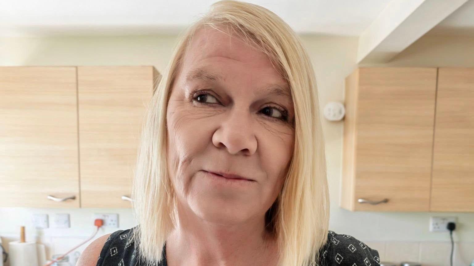 Jaqueline Watkins standing in front of cupboards. She is looking off to the side of the picture. She has shoulder-length blonde hair, with a side fringe.