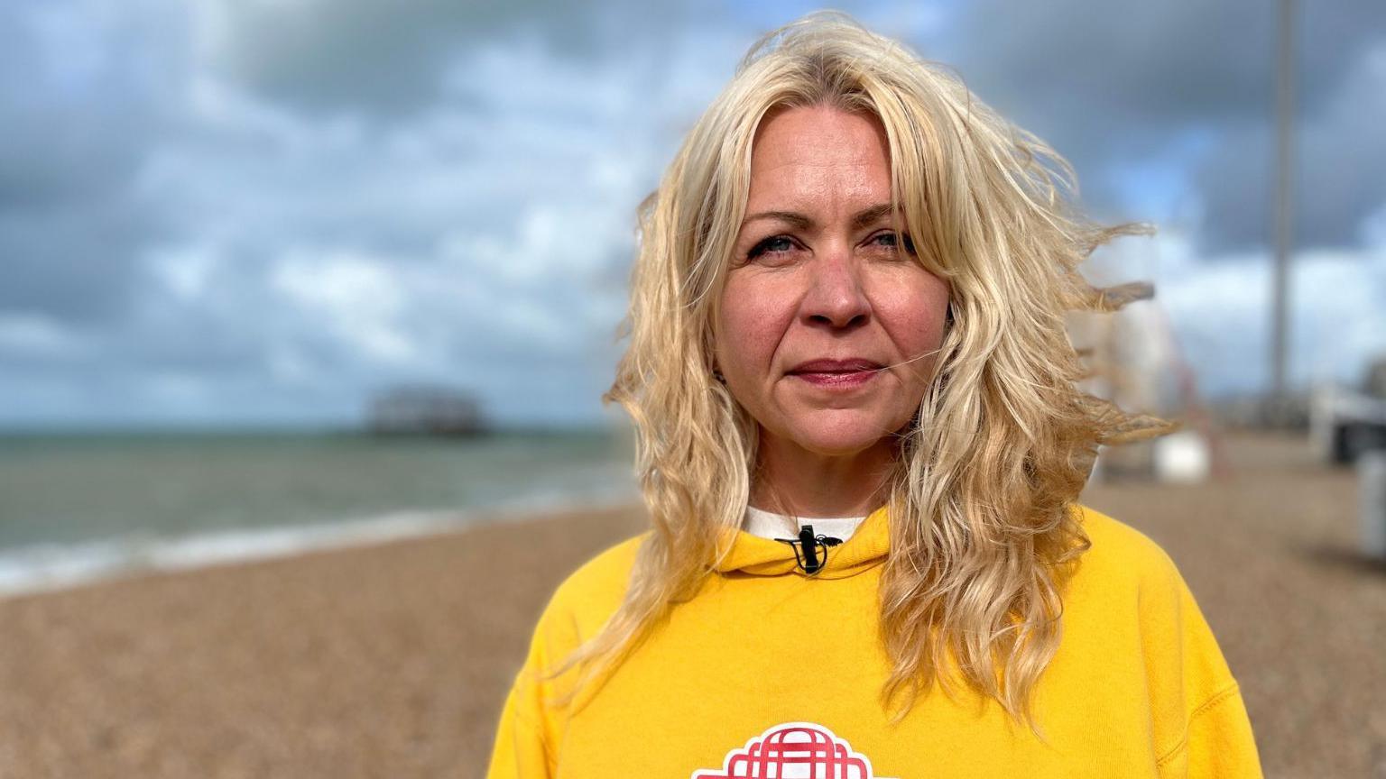 A woman with long blonde hair wearing a yellow hoodie with a red image of Brighton's West Pier. She is stood looking into the camera while stood on the pebbles of Brighton beach