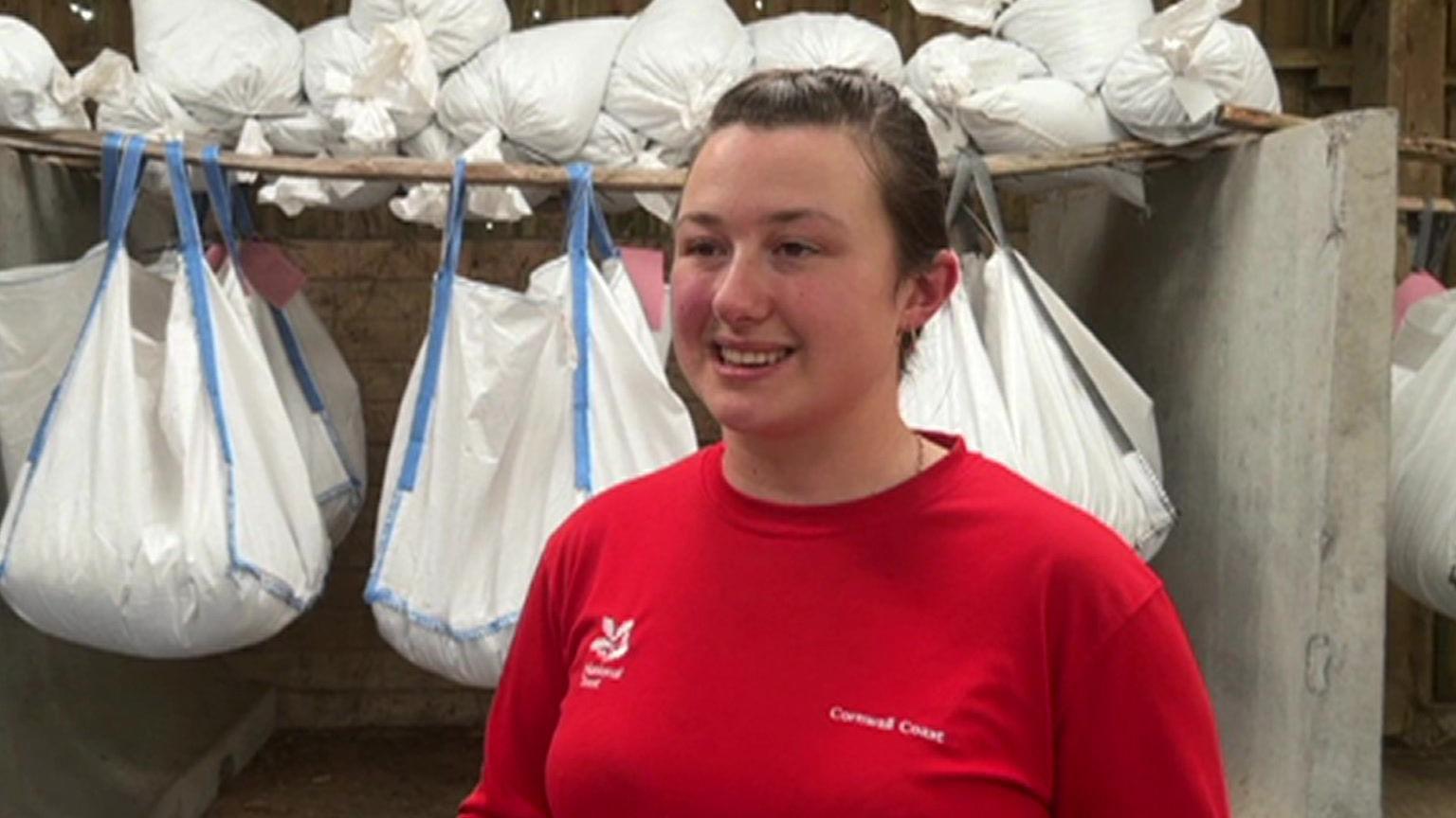 A photo of a woman called Fern. She is stood in the middle of the photo, surrounded by white and blue bags hung up behind her which contain seeds. Fern's hair is plaited behind her. She is wearing a red t-shirt with National Trust written on it in white.