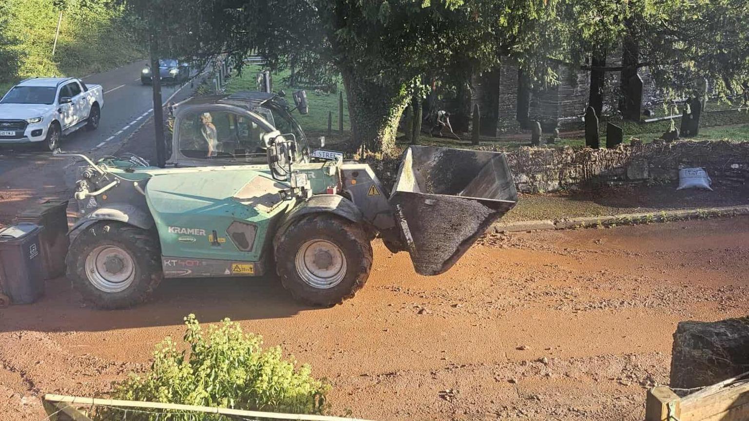 A large piece of machinery on a muddy road. It is green and there are cars travelling on the road behind. Behind the machinery are trees and a stone wall.