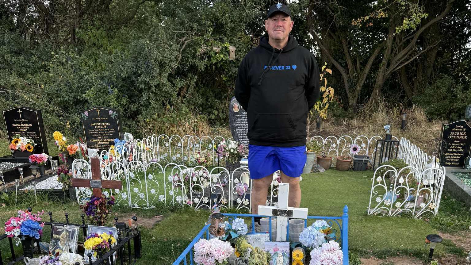 A man wearing a black hoodie and blue shorts stands behind a gravesite. The hoodie has the words 'Forever 23' written on the front.