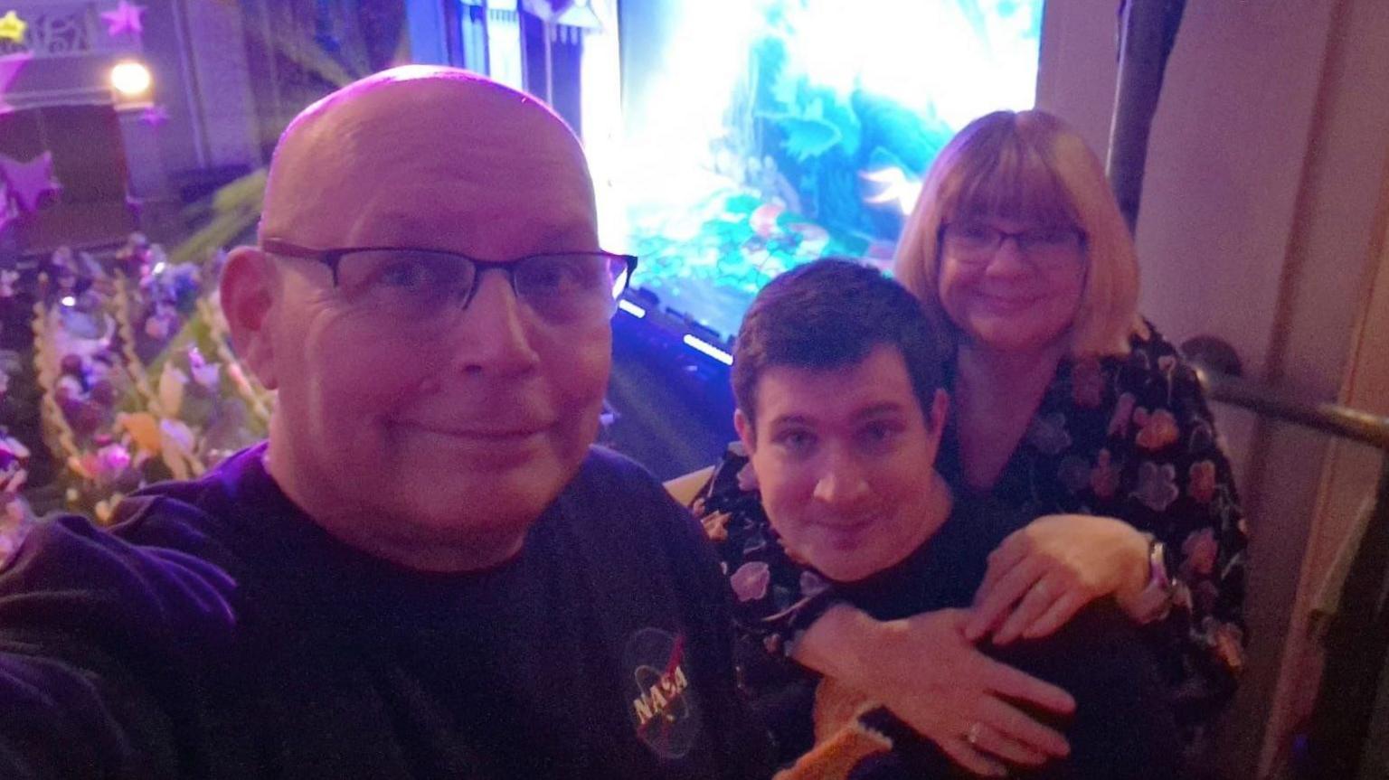 A selfie of a male and a female with their son in the middle. They are on a balcony in a theatre. The woman has short blonde hair and is wearing glasses. The father is bald and also wearing glasses. The son in the middle has short brown hair and is wearing a dark jumper. 