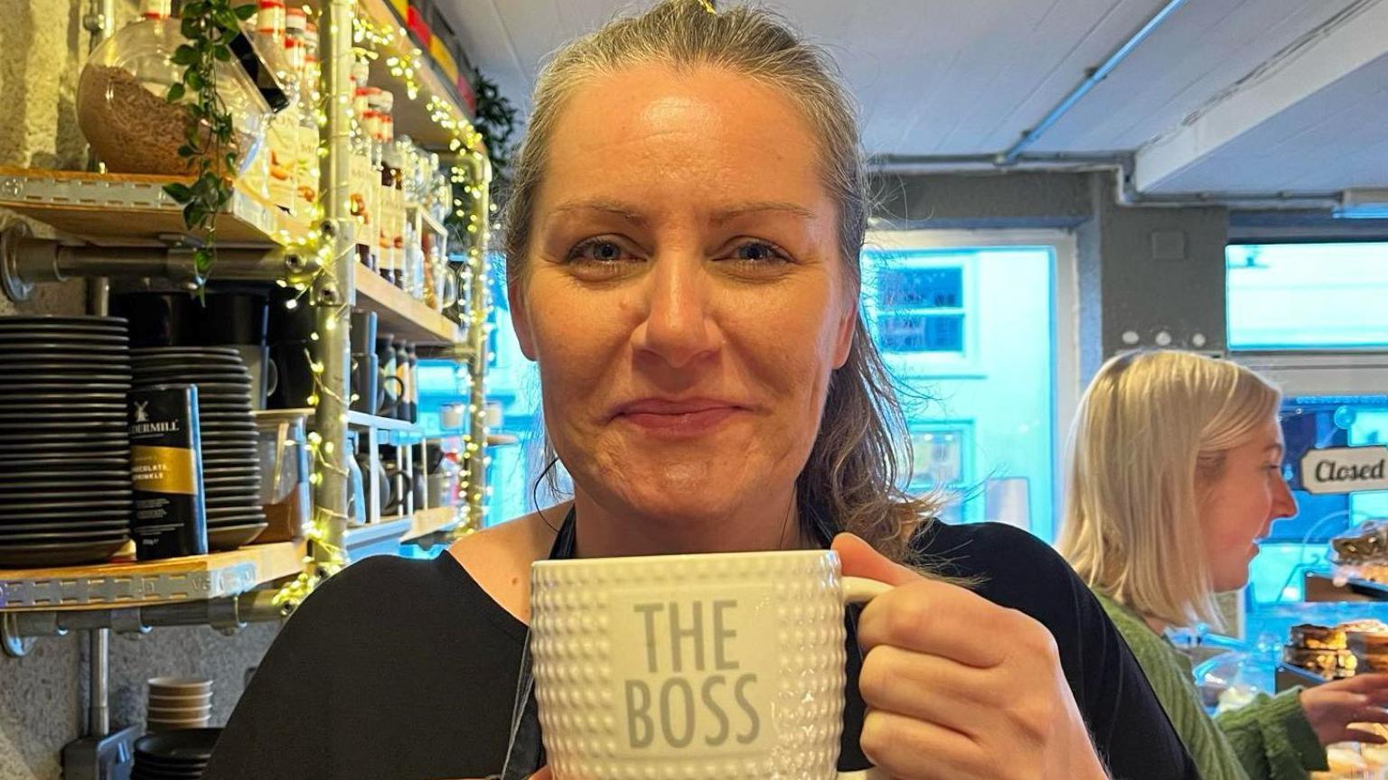A woman with grey hair in a ponytail holding a mug that reads the boss.