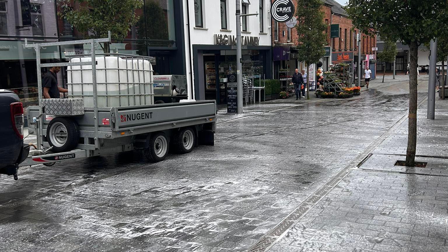 A black paved street with businesses down the side of it. There are suds on the ground as people clean up the slurry which was spread.