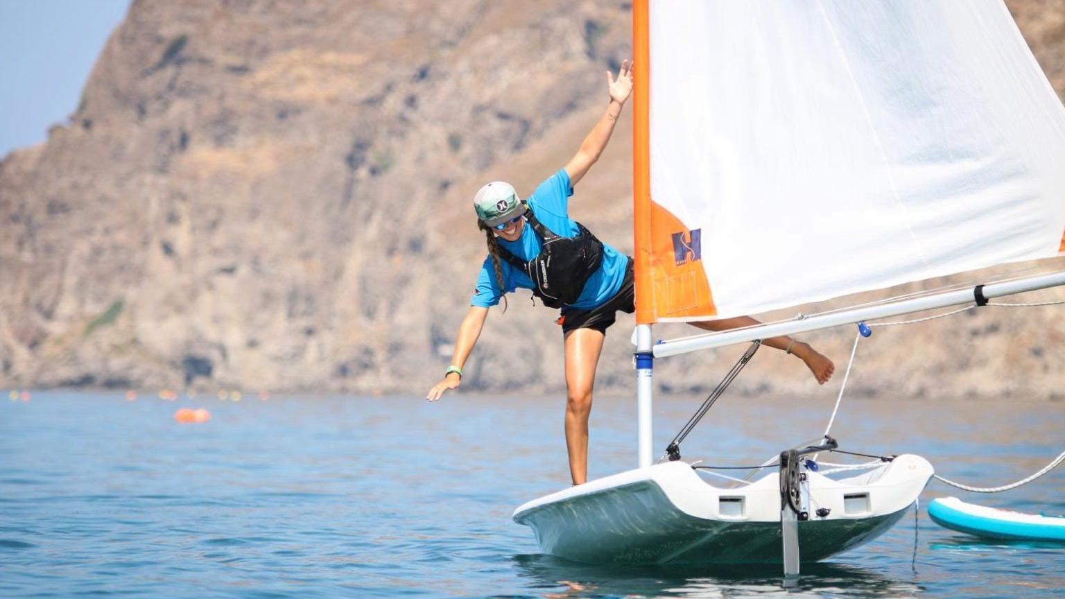 A woman wearing a blue tshirt, black shorts and a grey baseball hat stands on one leg on the front of a sailing dinghy. The sail is obscuring part of her other leg, which is barefoot. The sail is white and orange. She is smiling.