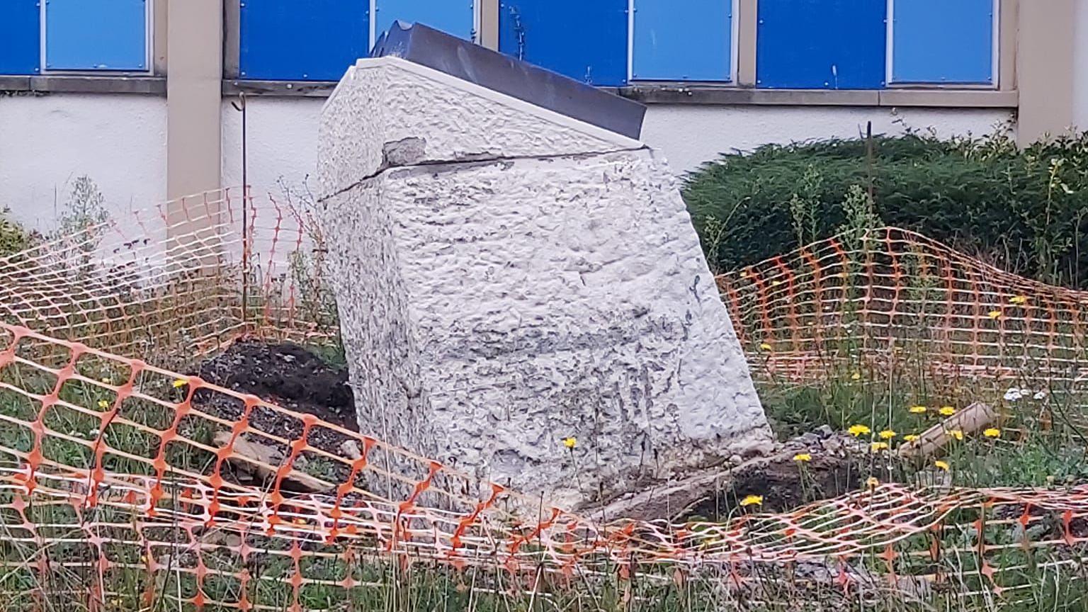 A tipped-over war memorial surrounded by orange plastic barrier fencing which is loose and untidy. There is soil uprooted at the bottom of the concrete base and there appears to be some chipped damage on the stone plinth.