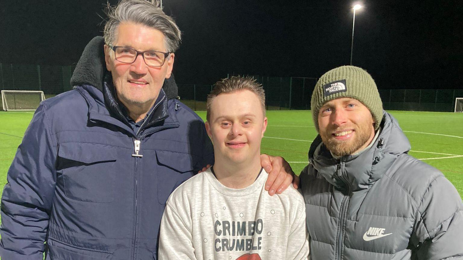 Luton Town's Mick Harford and Jordan Clark standing either side of James Forrester, who used to attend Redborne Upper School in Bedfordshire and now helps with the coaching. The three are smiling to camera and are on an astro turf pitch which is lit by flood lights. 

