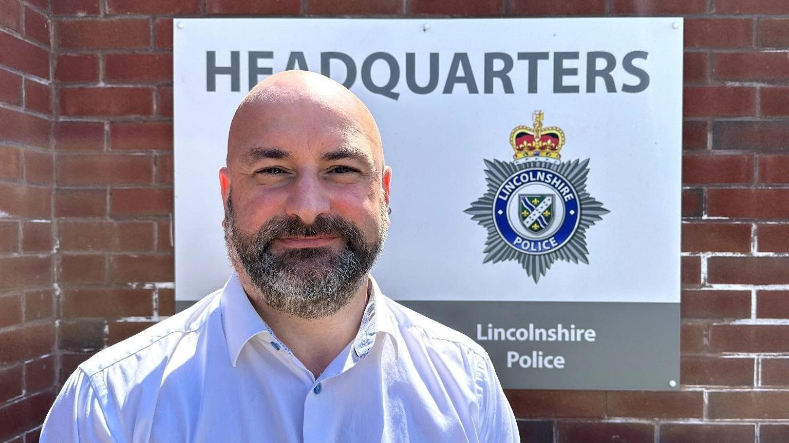 Mr Jones stands outside the Lincolnshire Police headquarters, he has a beard and is wearing a shirt with a collar.