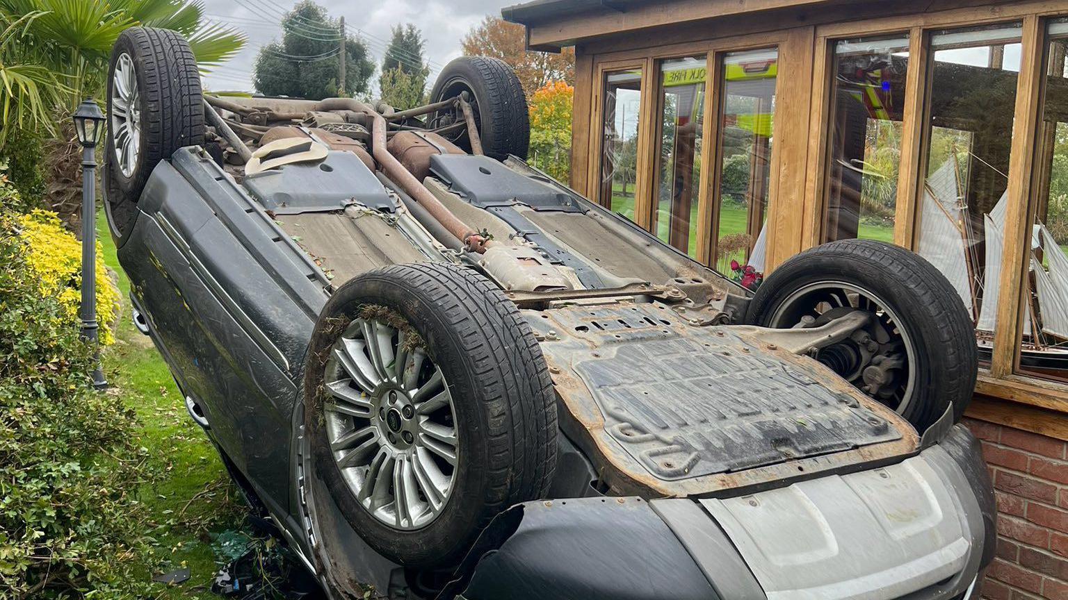 A silver car was turned on its roof and was in between a green hedge on the left and a building on the right which was made of red bricks and brown stained wood.