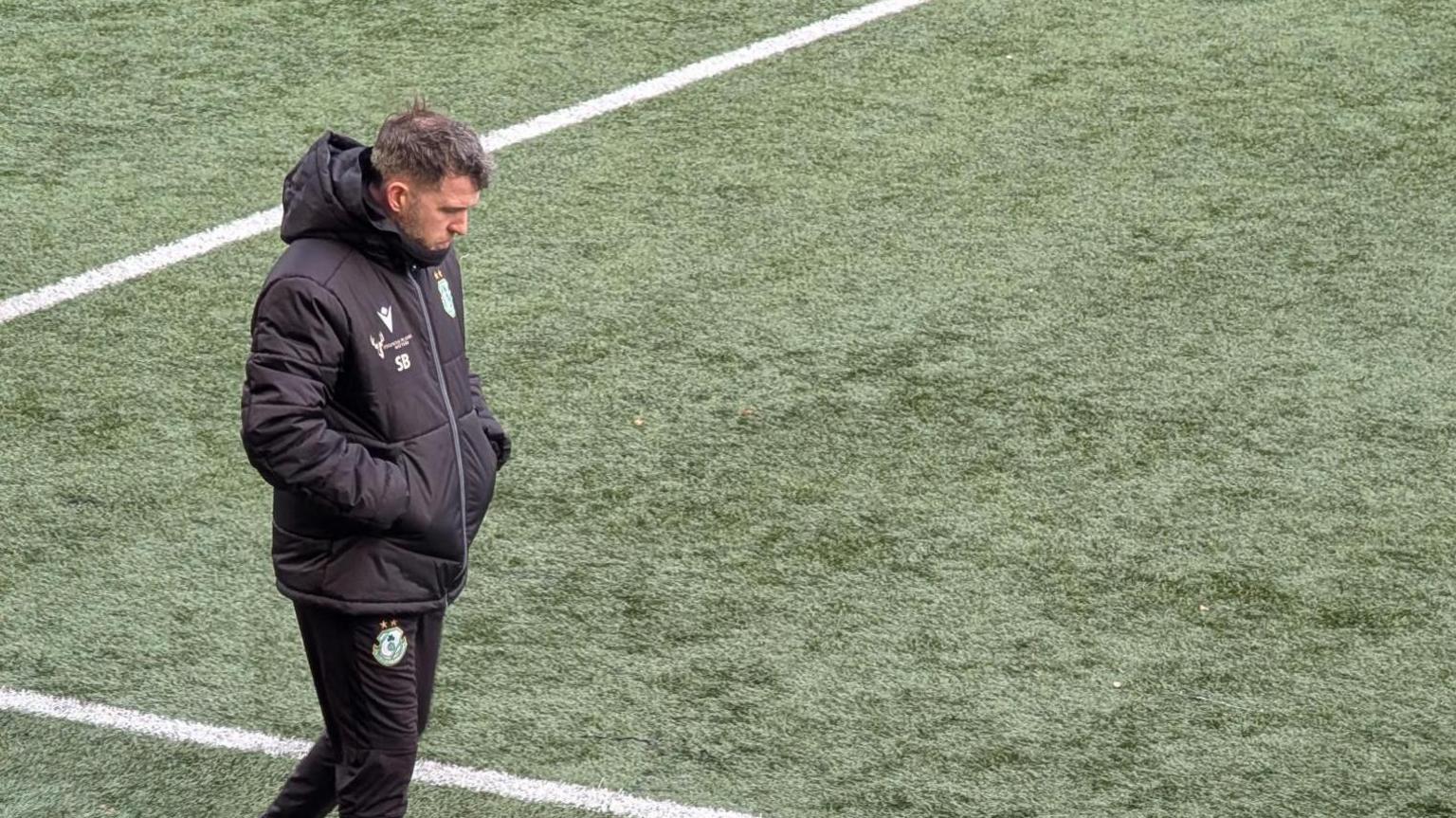 A man in a black coat and black trousers, both of which feature a green and white football crest, is walking along the side line of a football pitch