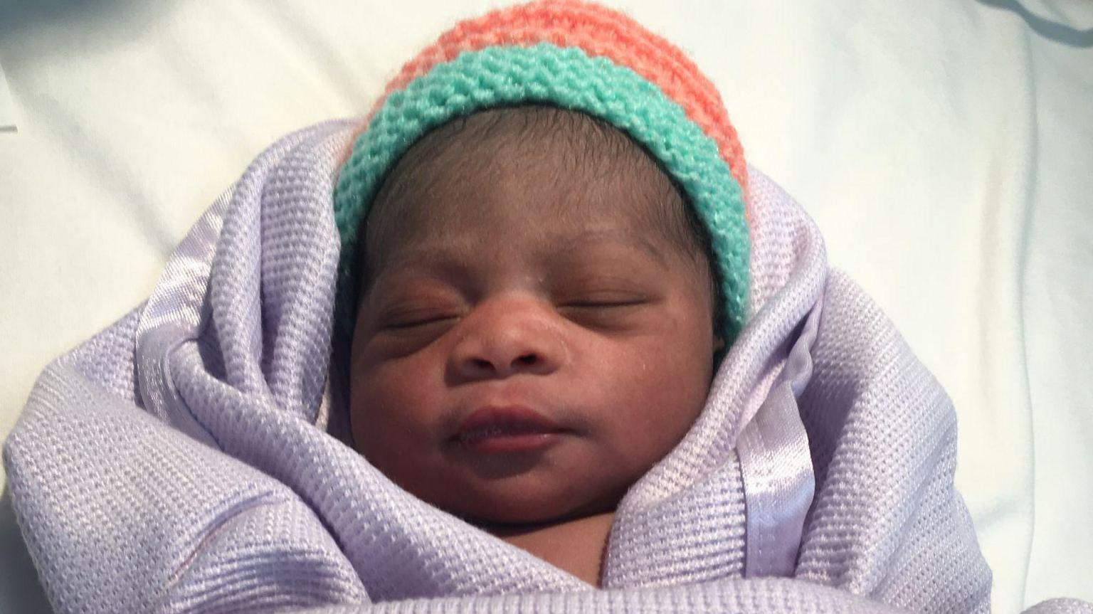 A new born baby boy wrapped in a lilac blanket and wearing a knitted hat with green and coral stripes.