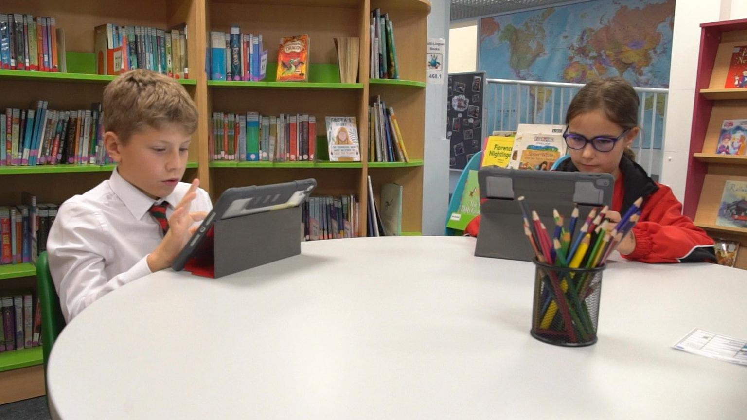 Two Jersey school pupils - a boy and a girl - sit at a desk in a library and look at computer tablets. The boy is wearing a white shirt and a red and green tie. The girl is wearing blue-rimmed glasses and a red coat. A pot full of colouring pencils is on the desk. A couple of bookshelves filled with children's books are behind the pupils.