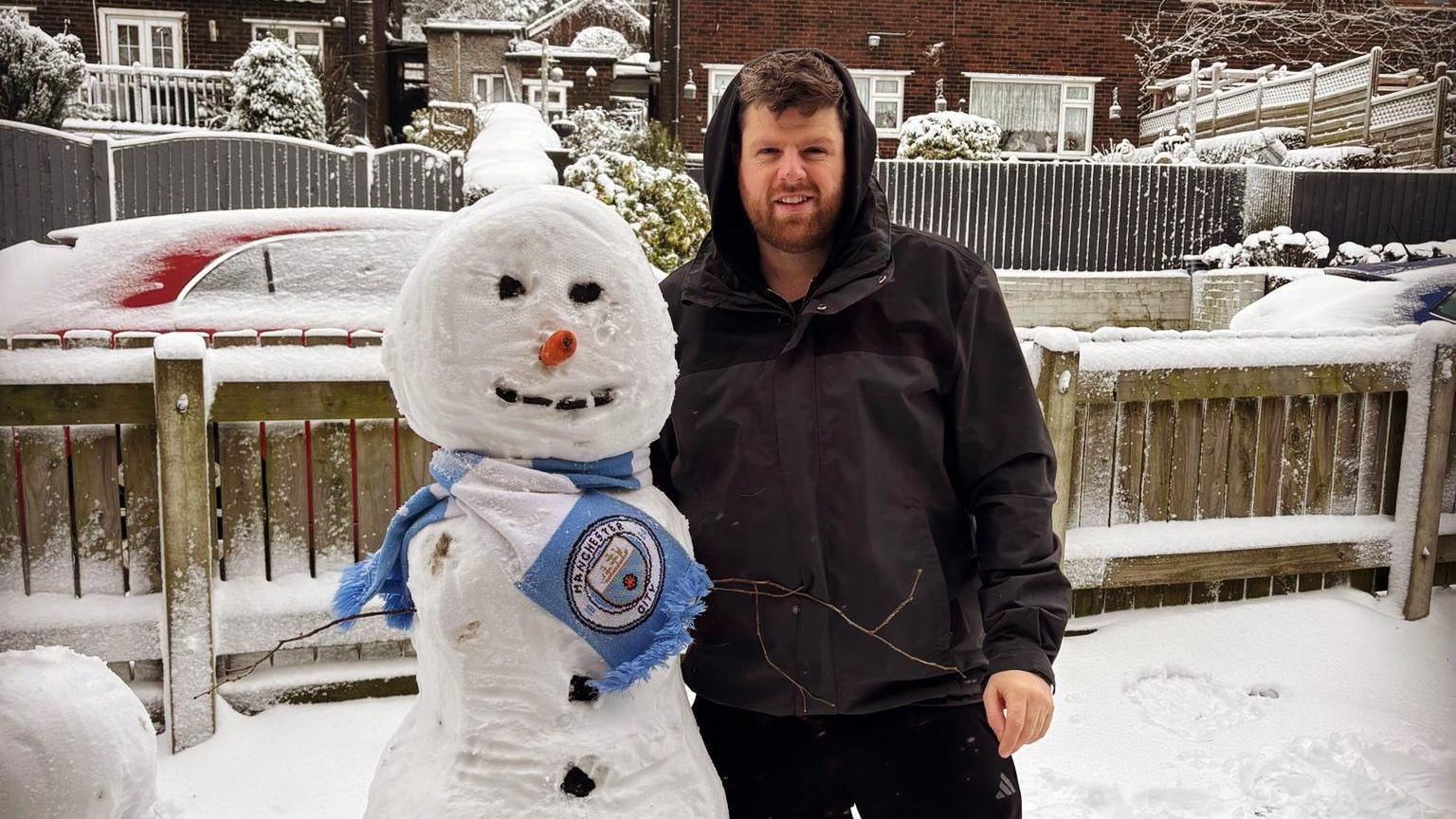 Tom McKeown stands next to his snowman, which is wearing a light blue Manchester City scarf, in his snow-covered garden. 