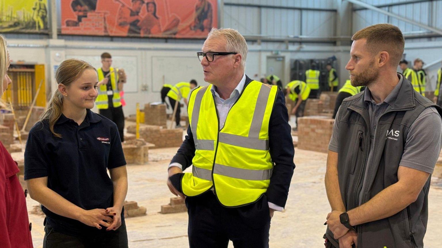 A man in the middle has white hair and glasses. Two people on either side of him are looking towards him. People in hi-vis jackets are working near bricks in the background.