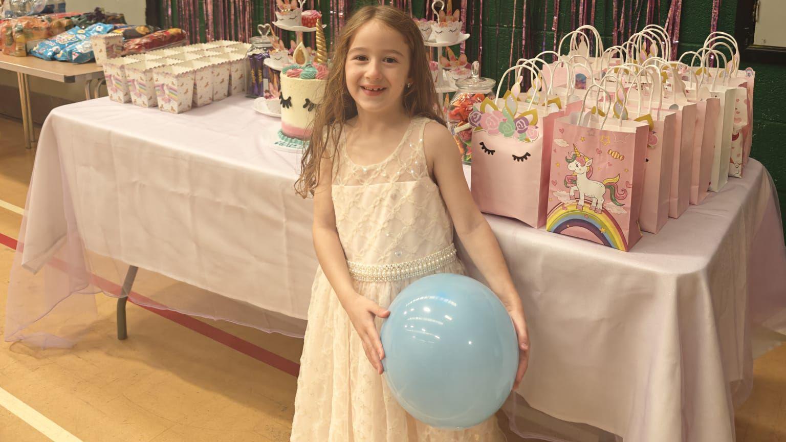 A little girl is standing in a village hall in front of a decorated table filled with party bags, a unicorn-themed birthday cake and smaller unicorn cakes on a cake stand. The girl is wearing a long white dress and is holding a light blue balloon. There is another table filled with presents in the background.