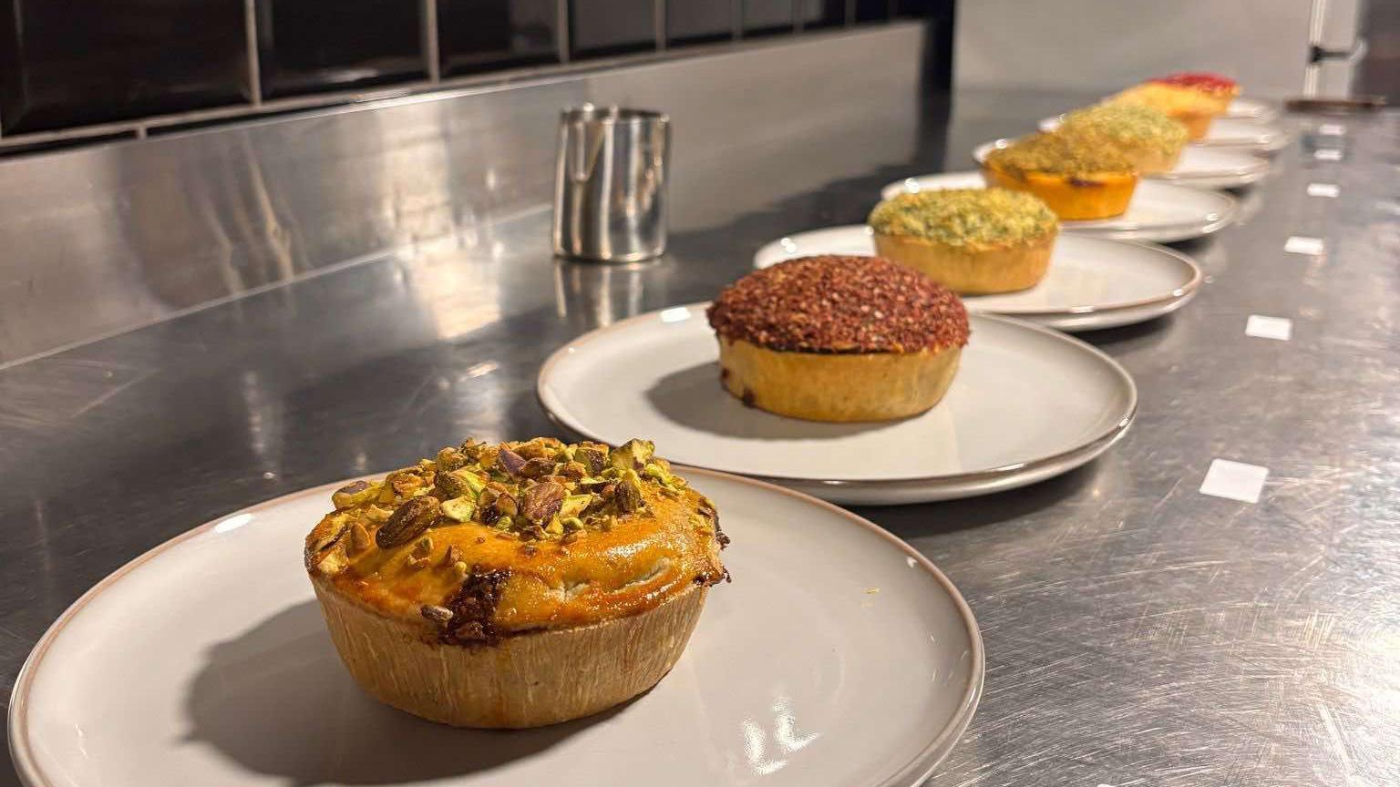 A row of different-flavoured pies lined up on white plates on a stainless steal kitchen workspace. 