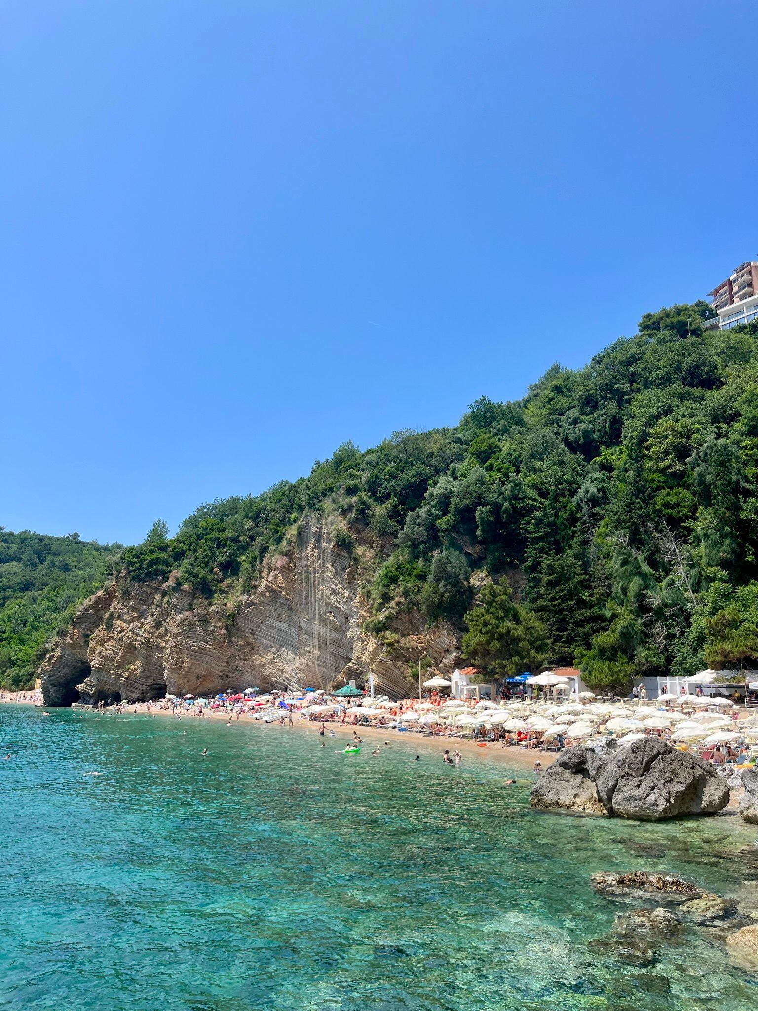 Imazh peizazhi i një plazhi në Mal të Zi. Ka një masë të madhe toke të lartë që shtrihet në det. Uji është blu i pastër dhe qielli është i kthjellët dhe me diell.