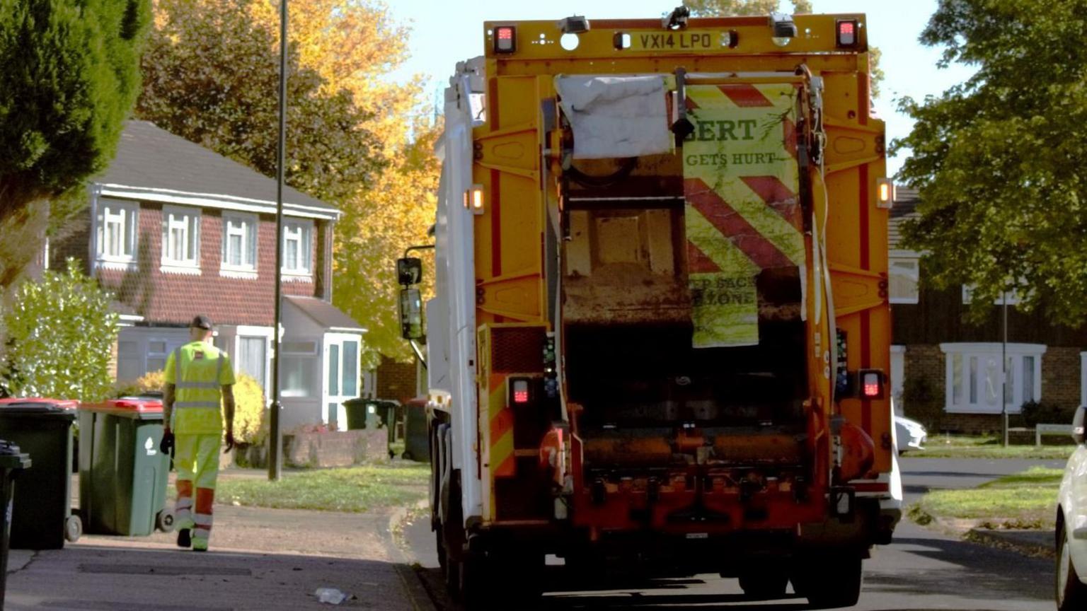 The back of a bin lorry. It is orange. A man in yellow overalls is walking on the pavement to the left of the lorry. There are green bins with red lids on the pavement. There are houses and a large green hedge on the left