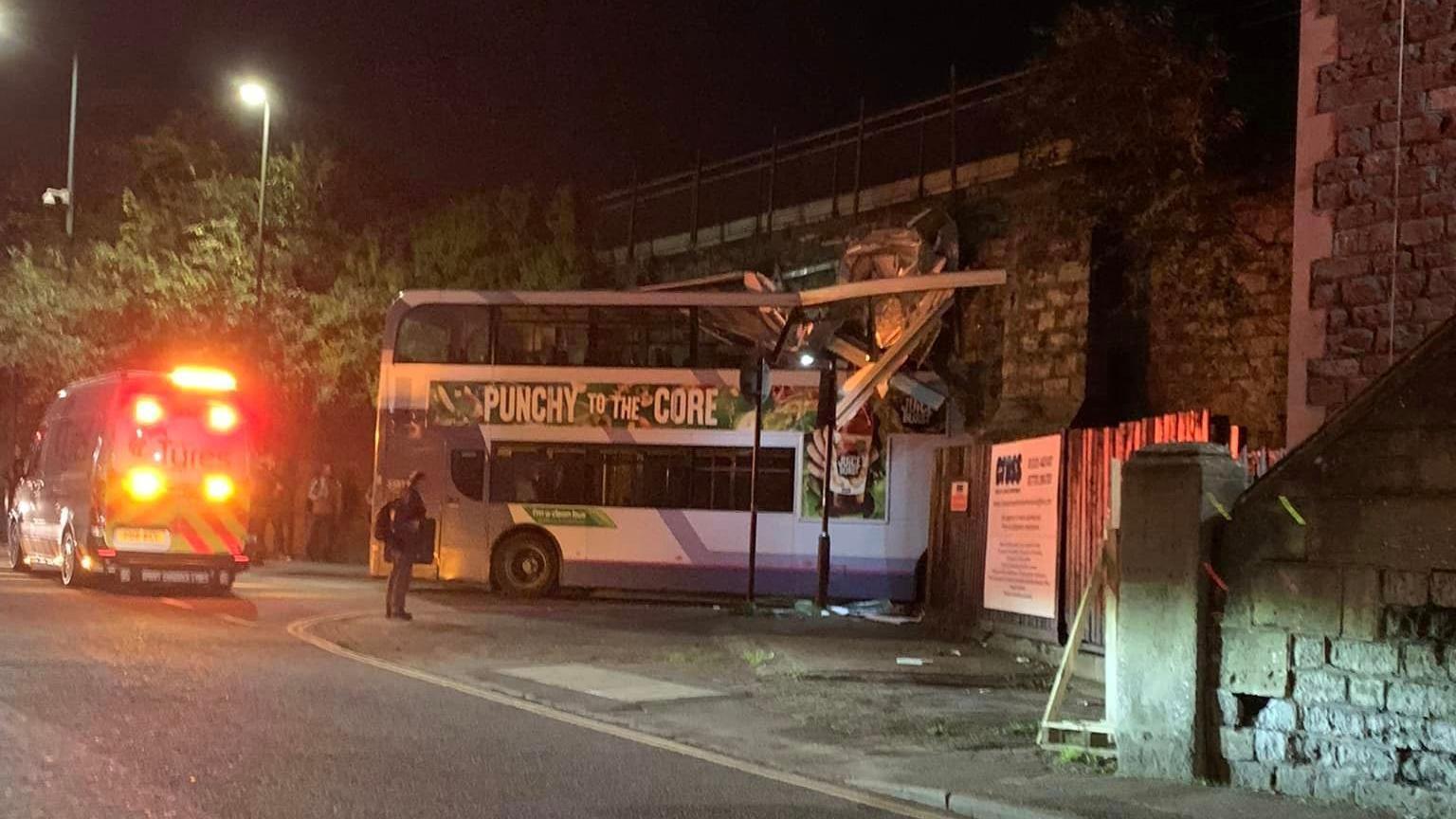 A double-decker bus lodged underneath a railway bridge. It is dark and there is a recovery vehicle parked behind it with its lights flashing. The front of the bus has been crumpled and torn apart significantly. 