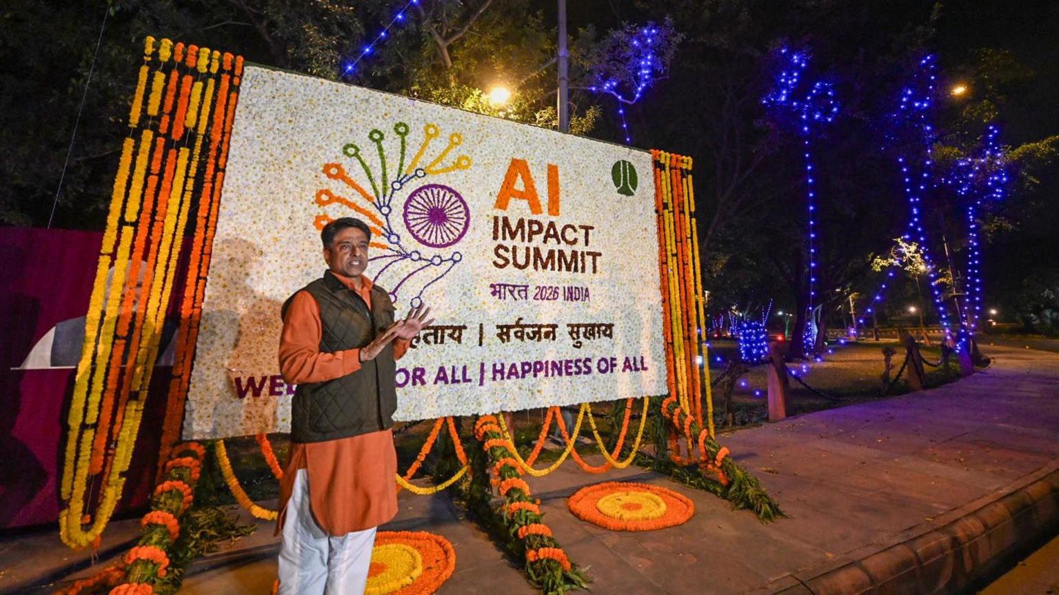 A man stands in front of an orange and white sign which reads "AI impact summit"