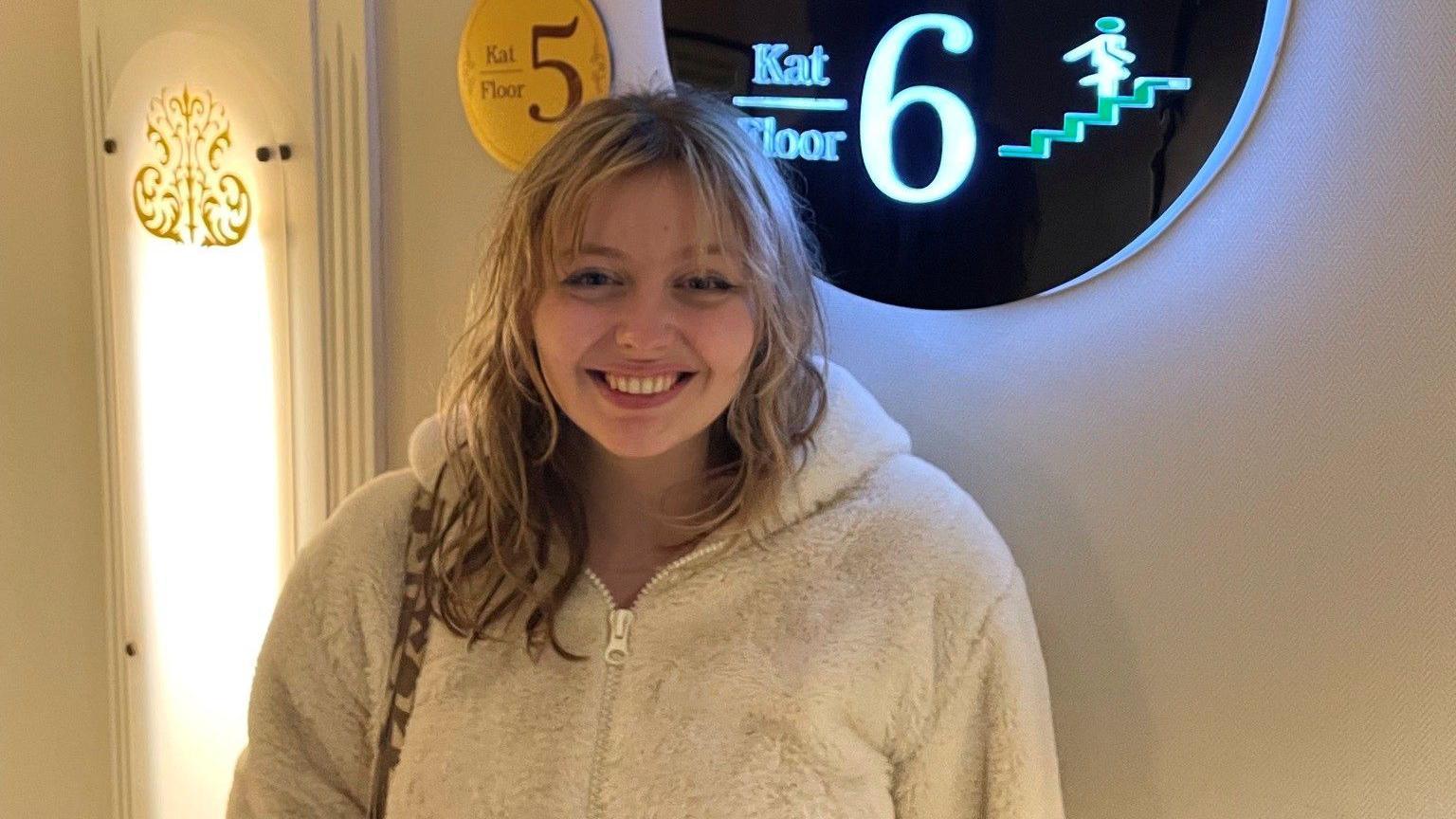 Kat Nellist is smiling at the camera while wearing a fluffy white jacket that has been zipped up to her neck. She has shoulder-length wavy blonde hair and curtain bangs. She is standing in front of a plain white wall with a neon sign behind her