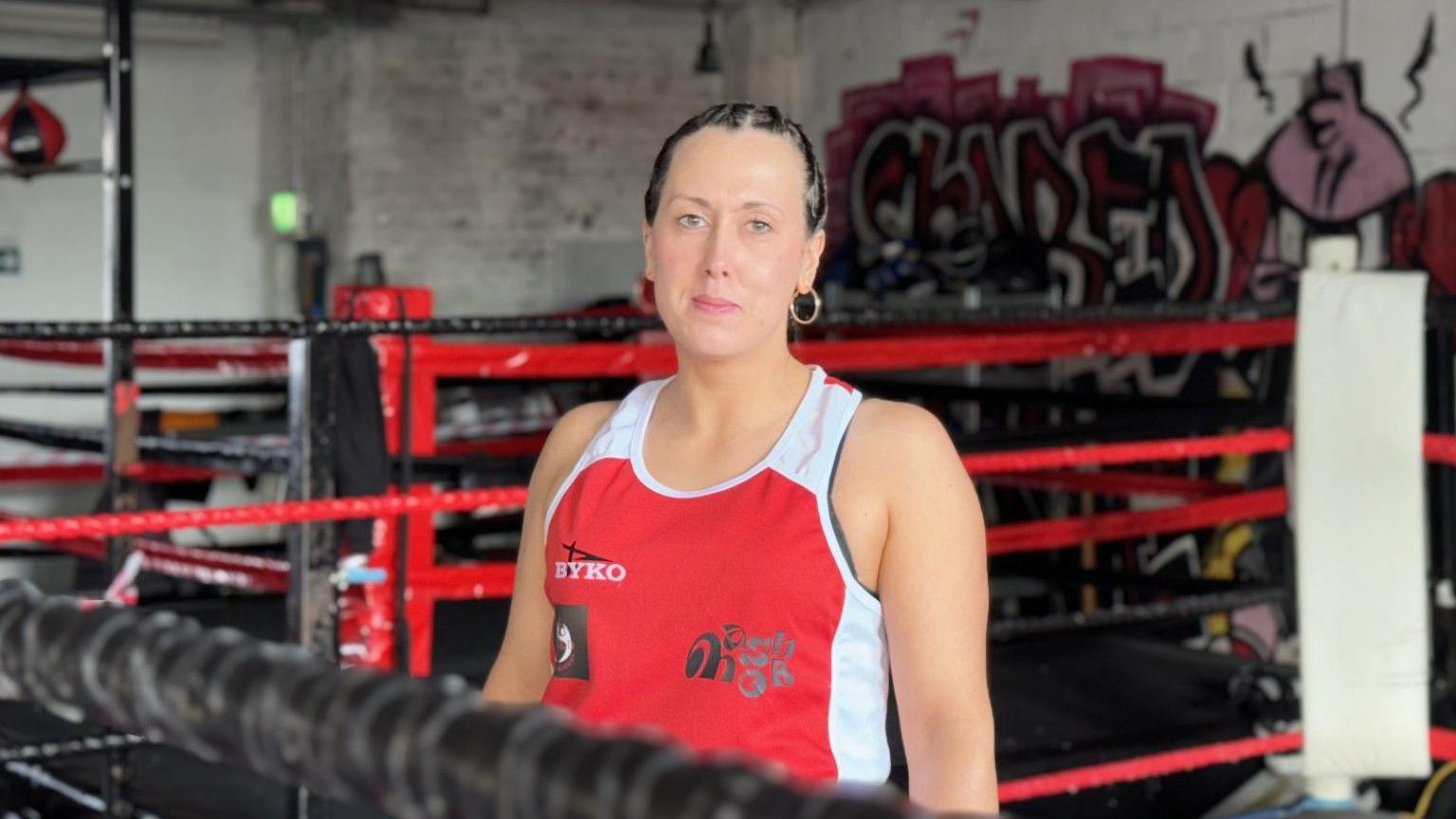 Leah Djalo looks over the top of a boxing ring rope - she has a red and white sleeveless boxing top on and she has black hair in a pony tail and a gold hoop earring is visible from her left ear.