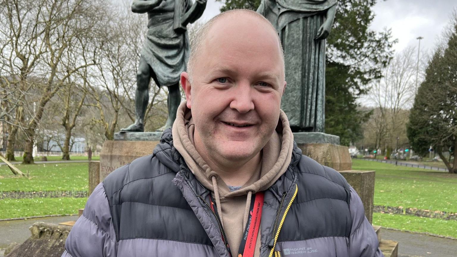 Prif weithredwr Menter Iaith Rhondda Cynon Taf, Osian Rowlands, o flaen cofeb Evan James a James James ym Mharc Ynysangharad, Pontypridd