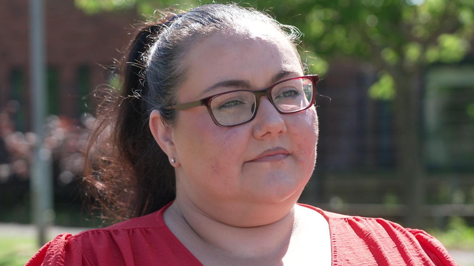 A woman is standing outside. her brown hair is pulled back in a ponytail. She wears red glasses and has a pink top on.