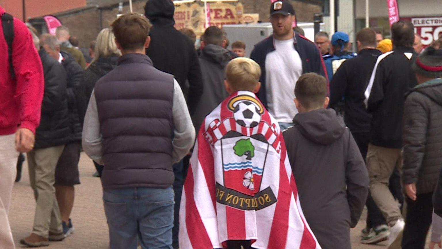 Southampton FC supporters outside the stadium. Three young fans walk together, one draped in the Southampton FC flag.