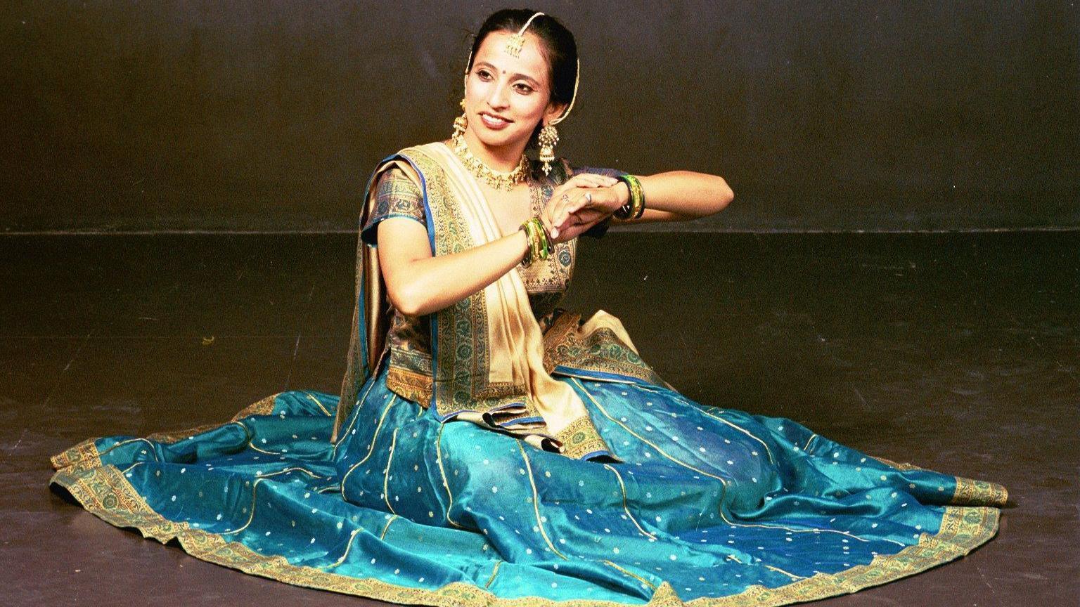 Urja Desai Thakore in traditional sari, sitting on the floor, as part of a dance. The sari is gold and blue in colour, she has jewellery on her face and is wearing bangles. She is smiling and looking slightly away from the camera.