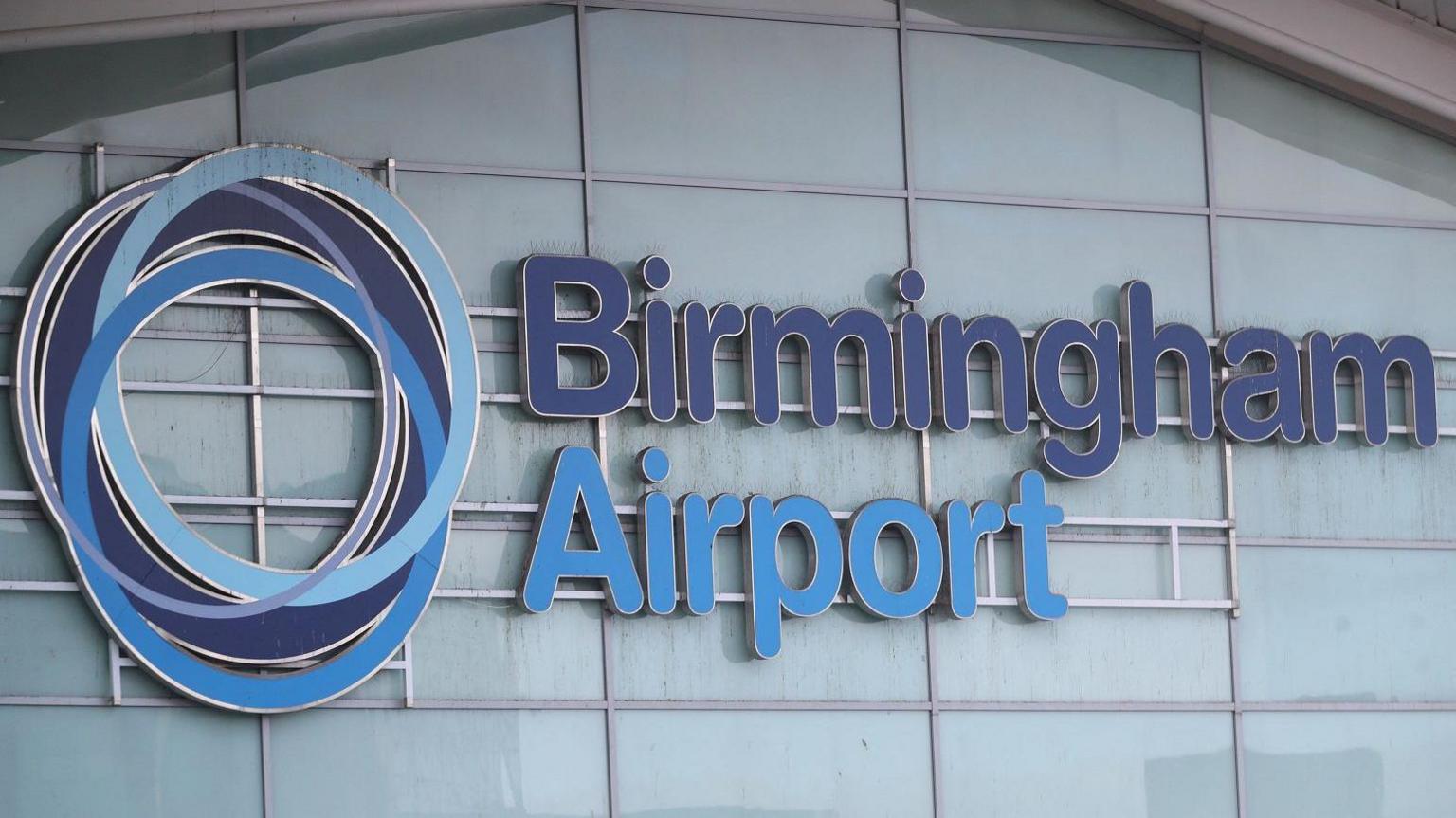 A sign saying "Birmingham Airport" in dark and light blue, on the side of a glass wall.