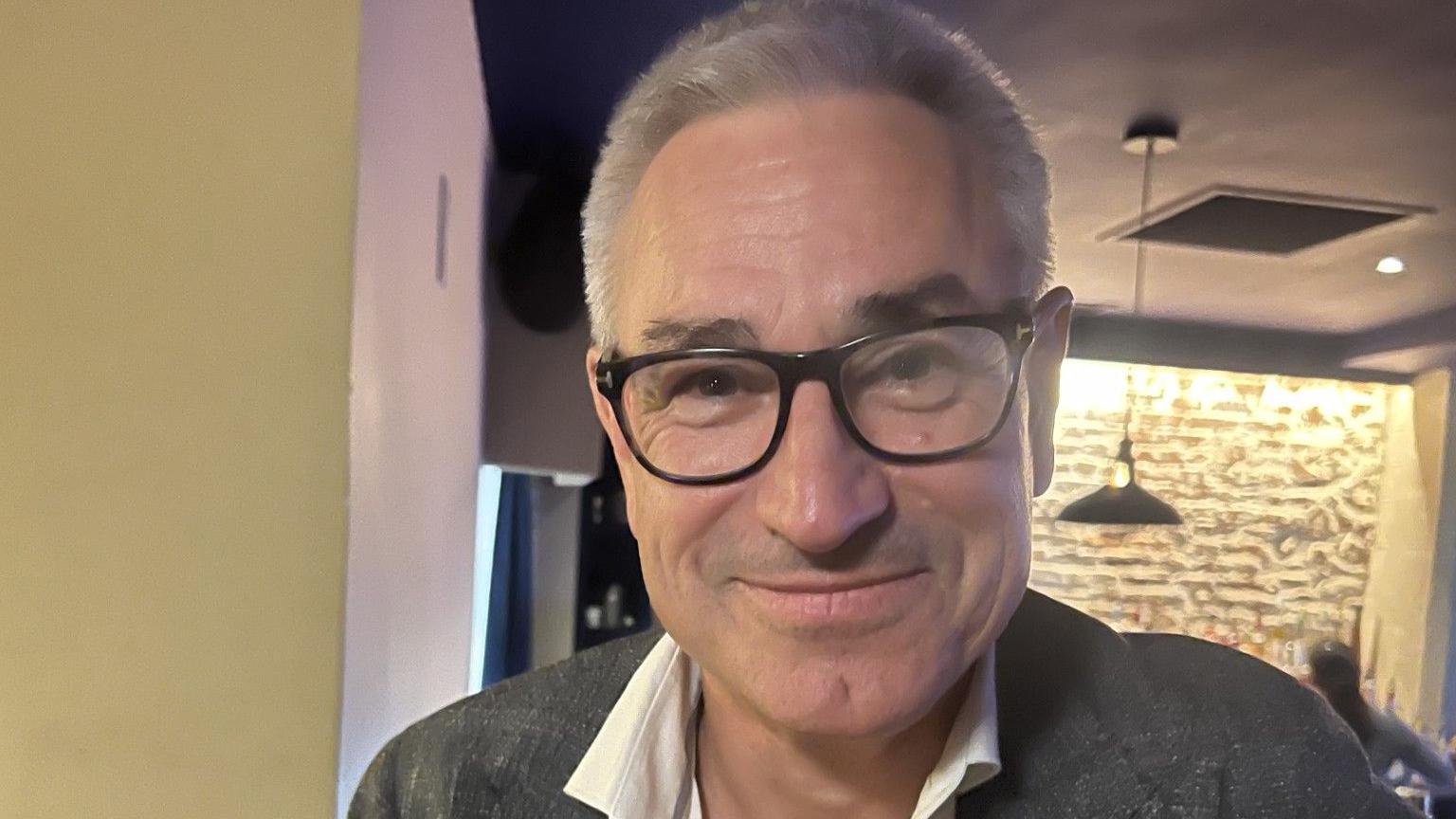 Gerald Voisin is pictured wearing a grey blazer, a white shirt and black-framed glasses. He has light grey hair and is smiling at the camera.