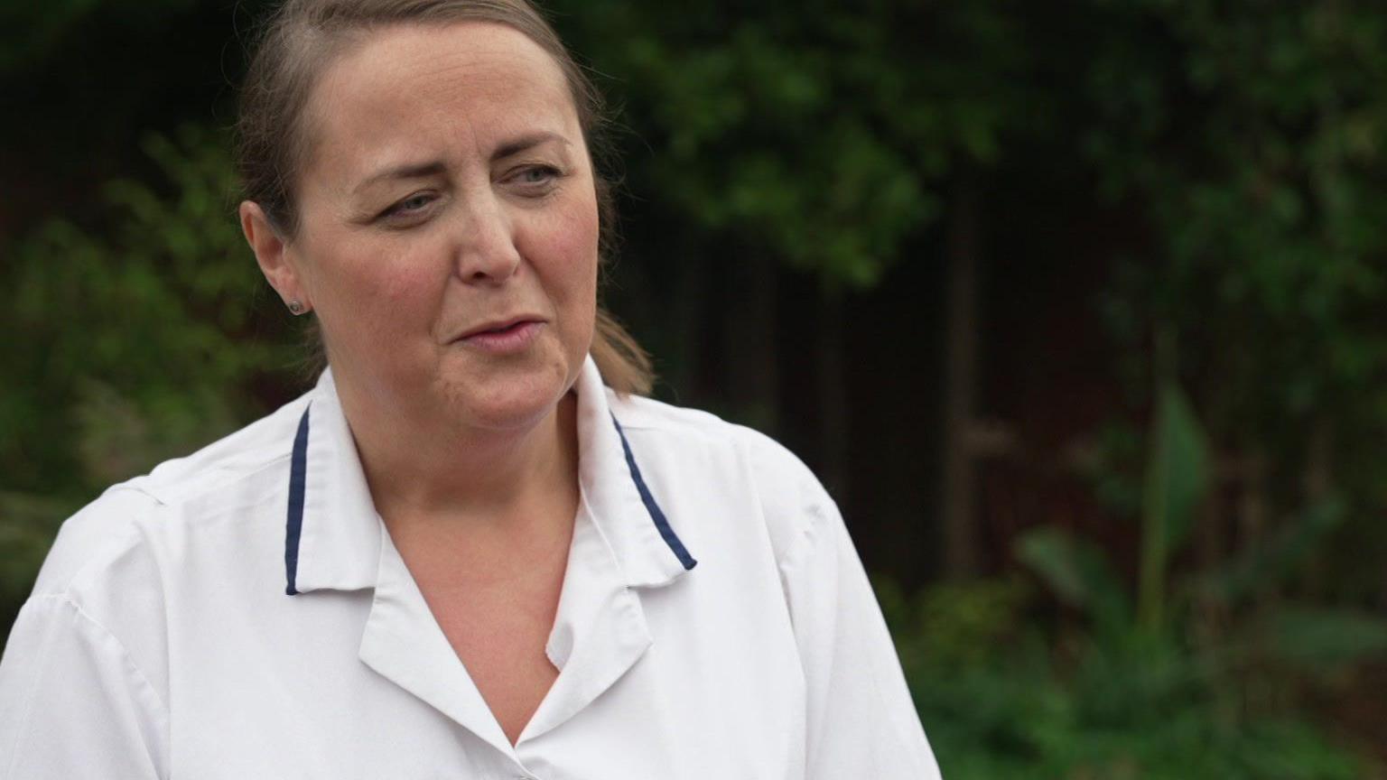 The picture shows a person wearing a white uniform with dark blue trim. The photo is taken outdoors, with greenery and trees in the background.
