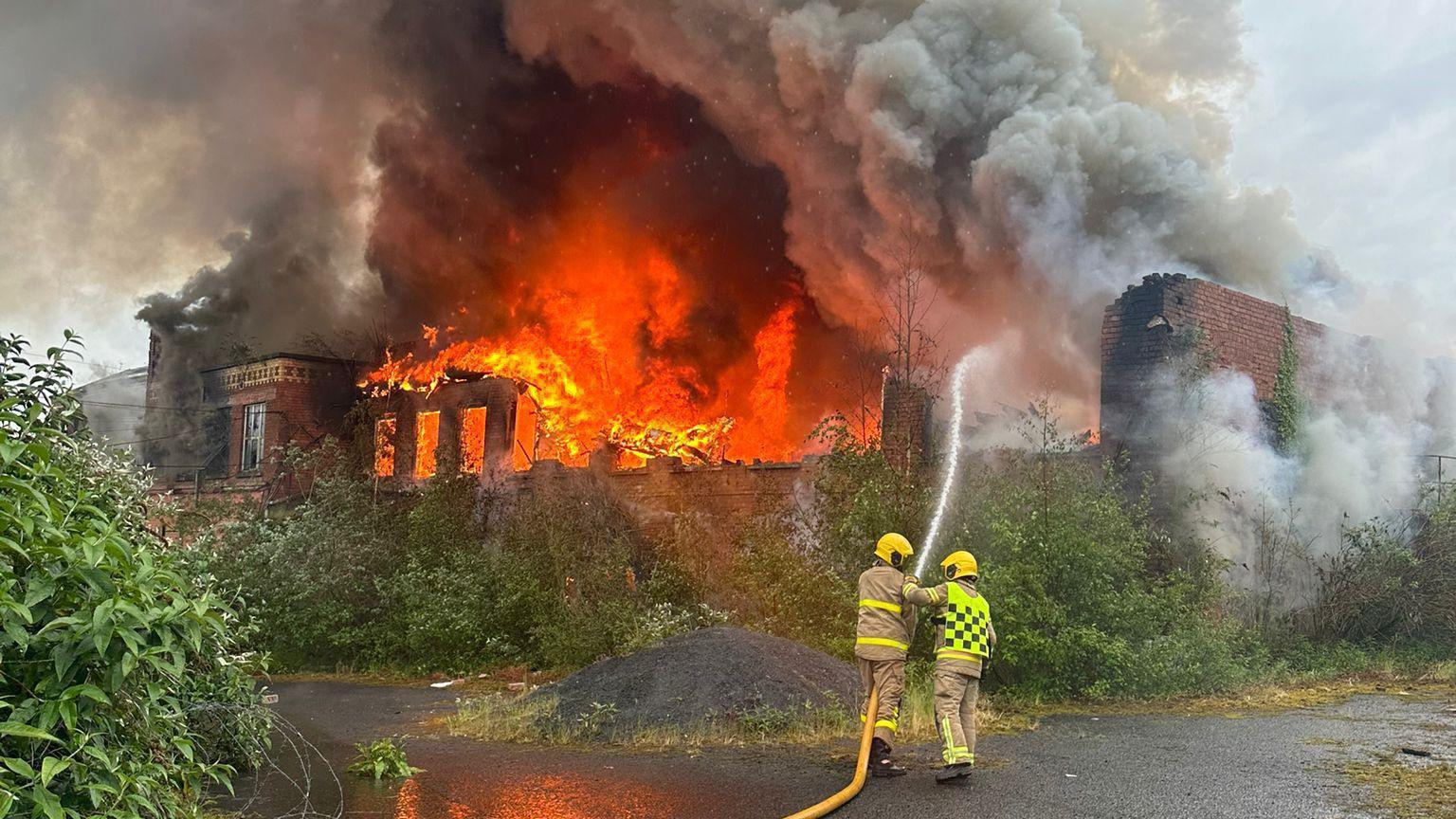 A deliberate fire at Hilden Mill in May 2024.  Two firefighters are using a hose to dampen a burning derelict building.  Flames have engulfed a large part of the brick structure and there are large plumes of grey smoke over the whole scene. 