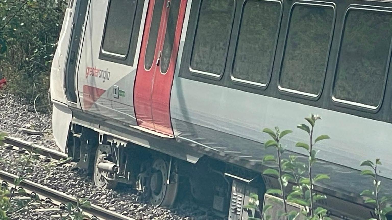 A close-up image of a Greater Anglia railway carriage. Its wheels have left the track and are resting in stones in between the metal lines. The carriage is upright, but at a slight angle.