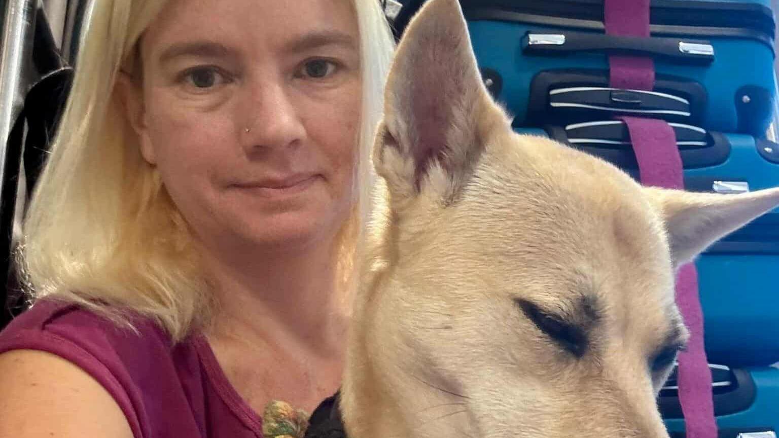 A close up of Naomi Rowan wearing a purple top and her dog Dusty in a hotel room. The pair take up most of the frame but you can still her pile of suitcases behind her on her left.