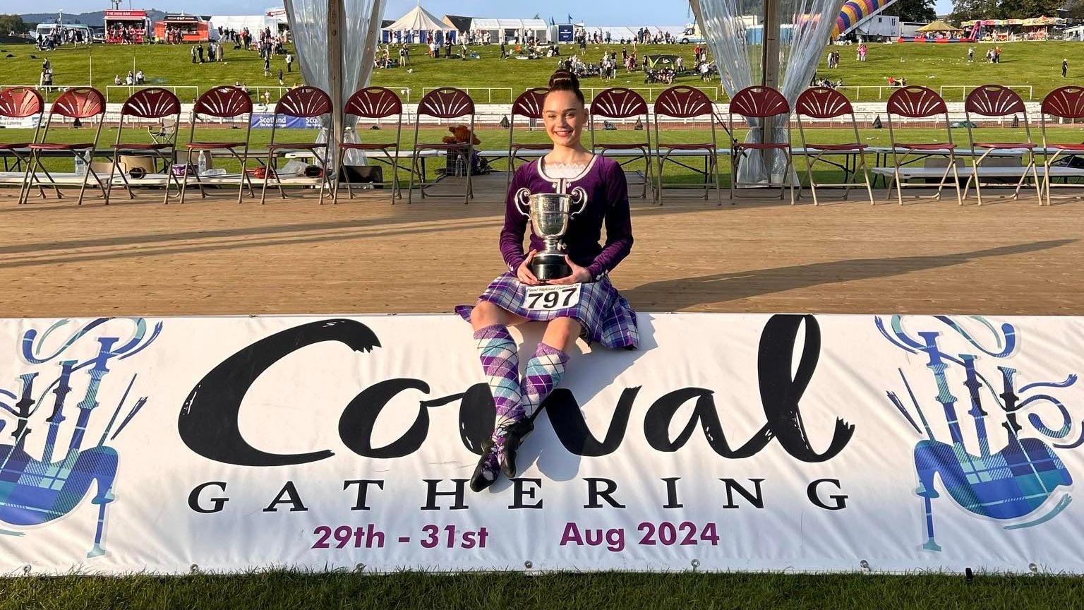The image shows a young girl, wearing a purple and white highland dance costume, sitting on a banner that reads "Cowal Gathering 29th-31st Aug 2024". She is holding a trophy.