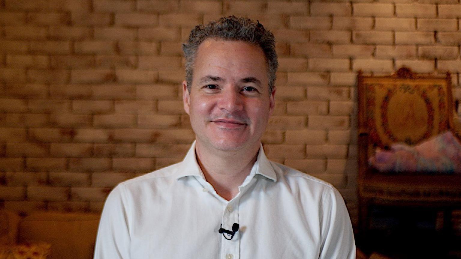 A man with grey hair and a white shirt looking at the camera. He is smiling and sat in front of a light brown brick wall. There is a small black microphone clipped to his shirt