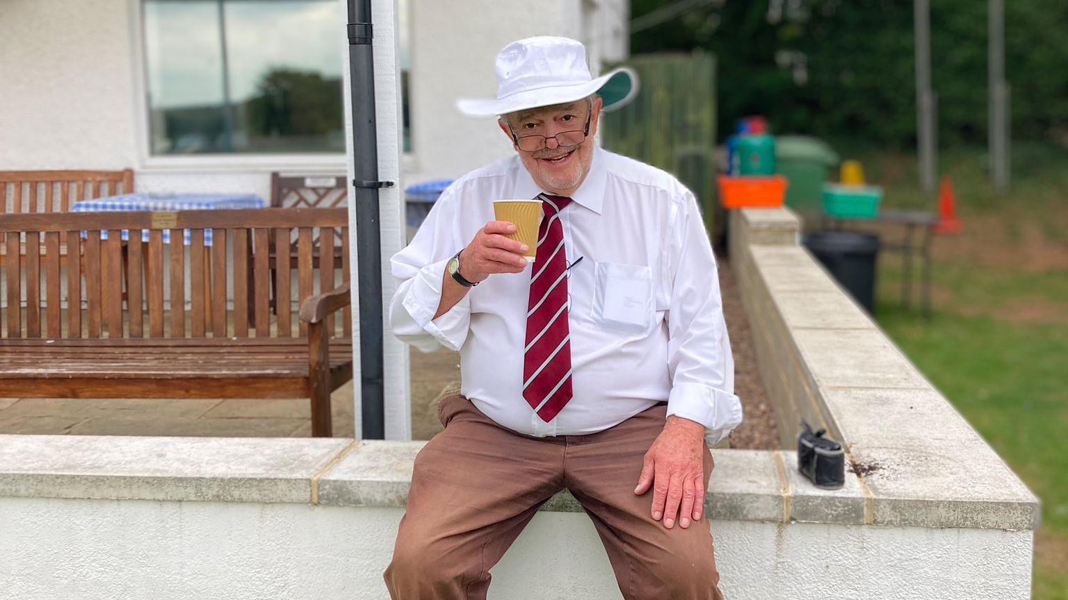 A man sitting on a wall holding a cup of tea, wearing a white shirt and red tie, a sun hat and brown trousers. 