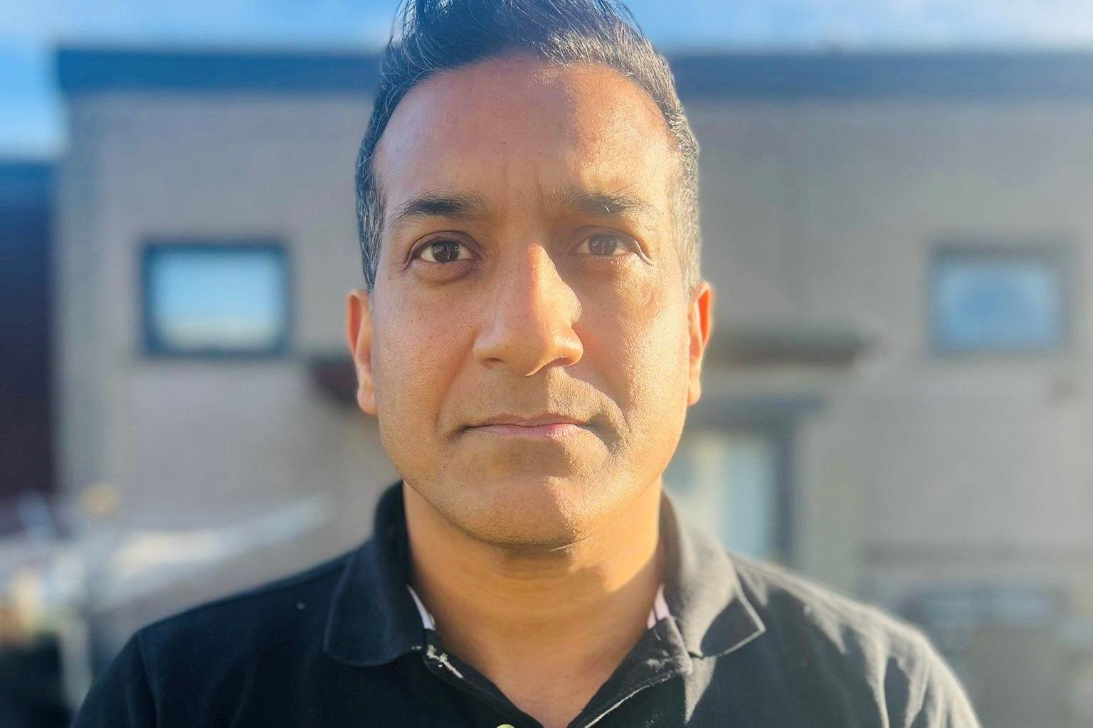 Man, Wilson Chowdhry, with black hair and black top, looking a camera, face lit up by sunlight, with building in background.