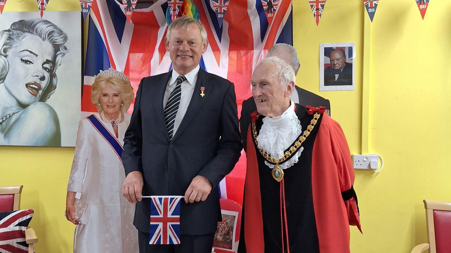 A smiling Martin Clunes standing next to the mayor of Ferndown, who is wearing his red ceremonial robes and chains. Clunes is wearing a suit and holding a small union flag in front of him. Behind him is a life-sized cardboard cut out of Queen Camilla and on the wall is a large portrait of Marilyn Monroe.