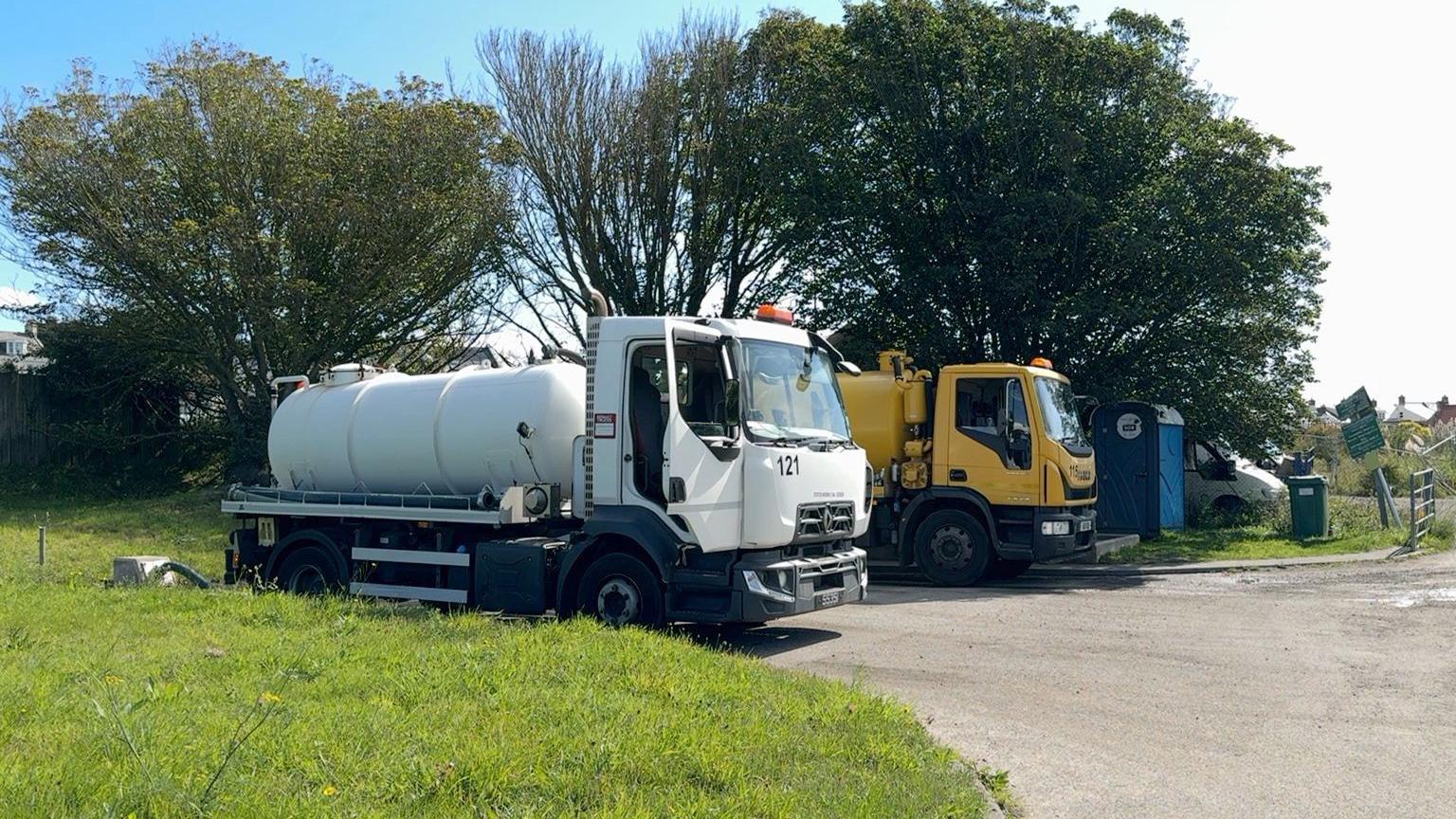 A white sewage cart lorry and a yellow sewage cart lorry in front of some trees. 